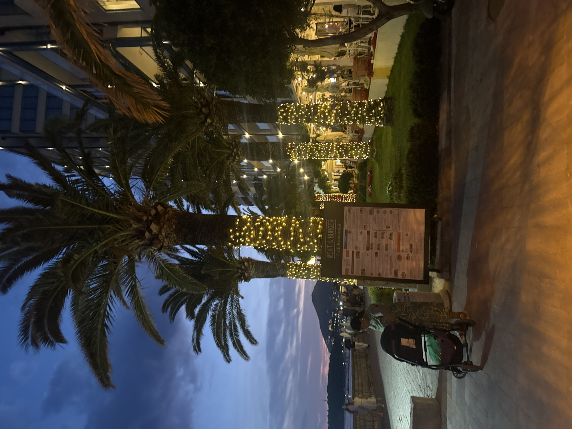 The image depicts a twilight scene along a waterfront promenade in Bečići, Montenegro, with a prominent "Meat & Burger Grill Restaurant" menu board. Several palm trees, adorned with warm-toned string lights, line the walkway. In the foreground, a woman is standing next to a stroller, appearing to be engaged with it. Behind her, other pedestrians are visible on the promenade, walking towards a body of water. In the background, a hill with scattered lights and a building are silhouetted against the darkening sky. The promenade is paved with cobblestones, and a strip of grass with bushes and potted plants is situated between the walkway and the restaurant area, which features outdoor seating with umbrellas. The overall atmosphere is tranquil and illuminated by the ambient light of the setting sun and artificial lighting.
