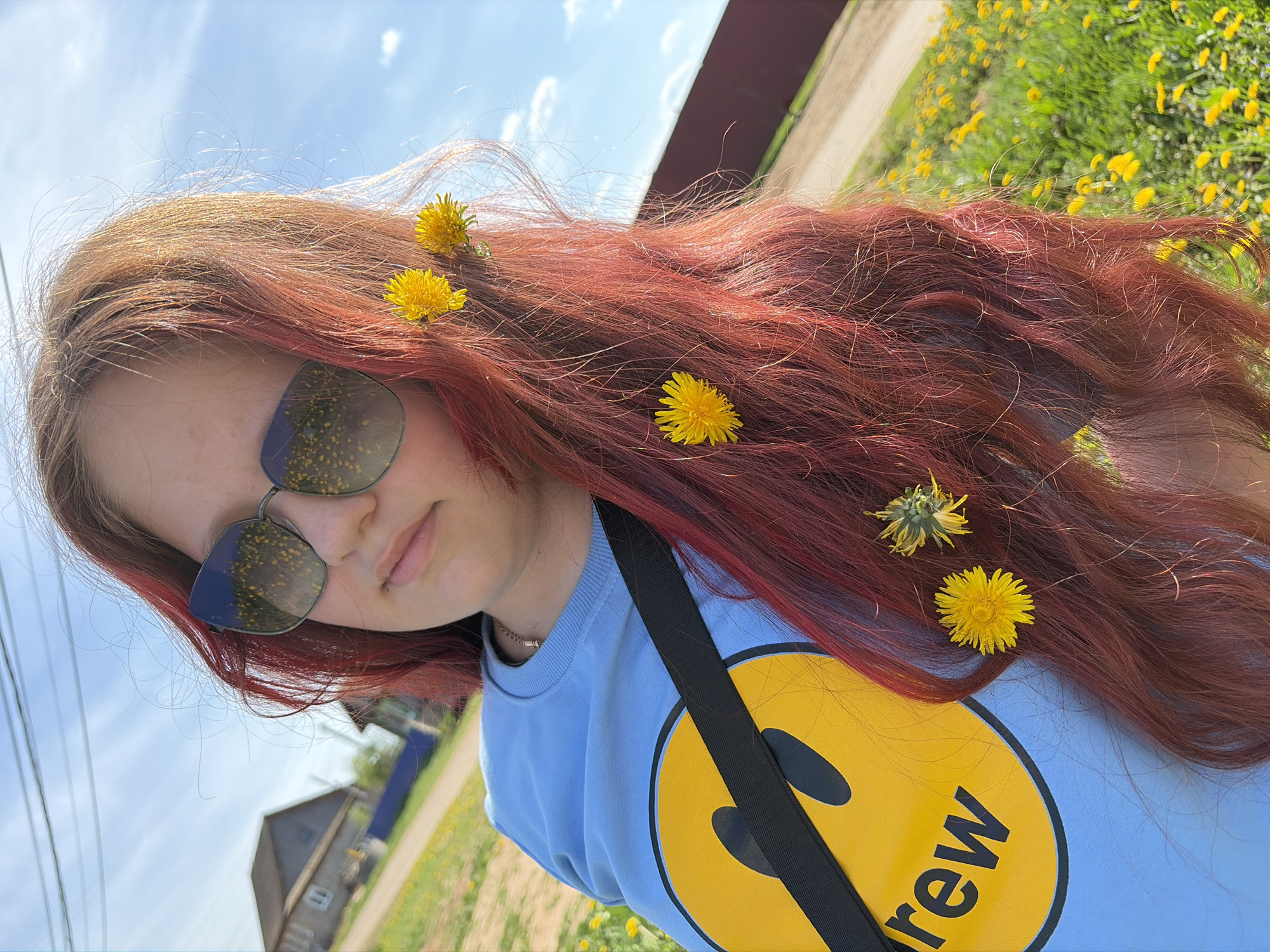 A young person with long red hair and sunglasses is posing in an outdoor setting. Yellow dandelions are woven into the person's hair. The person is wearing a light blue sweatshirt with a large yellow smiley face design and a black strap across the shoulder. In the background, there is a grassy area with more dandelions, a building, and a blue sky. While there is a city called Чумна in Russia, there is no information suggesting that is where the image was taken. Therefore, location cannot be confirmed.