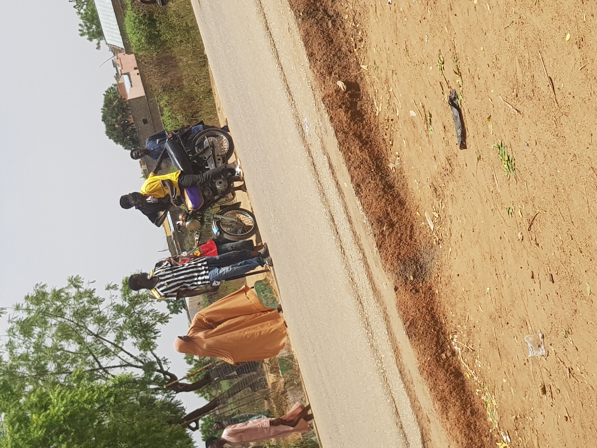 The image shows a street scene in Dabawa, Nigeria. Several people are visible on the side of a paved road. Two men are near motorcycles. One man wears a yellow shirt, and another wears a black and white striped shirt. A woman in a tan, full-body garment stands nearby. A tree with green foliage is visible to the left. The terrain beside the road is dry, brown soil with sparse vegetation and litter.