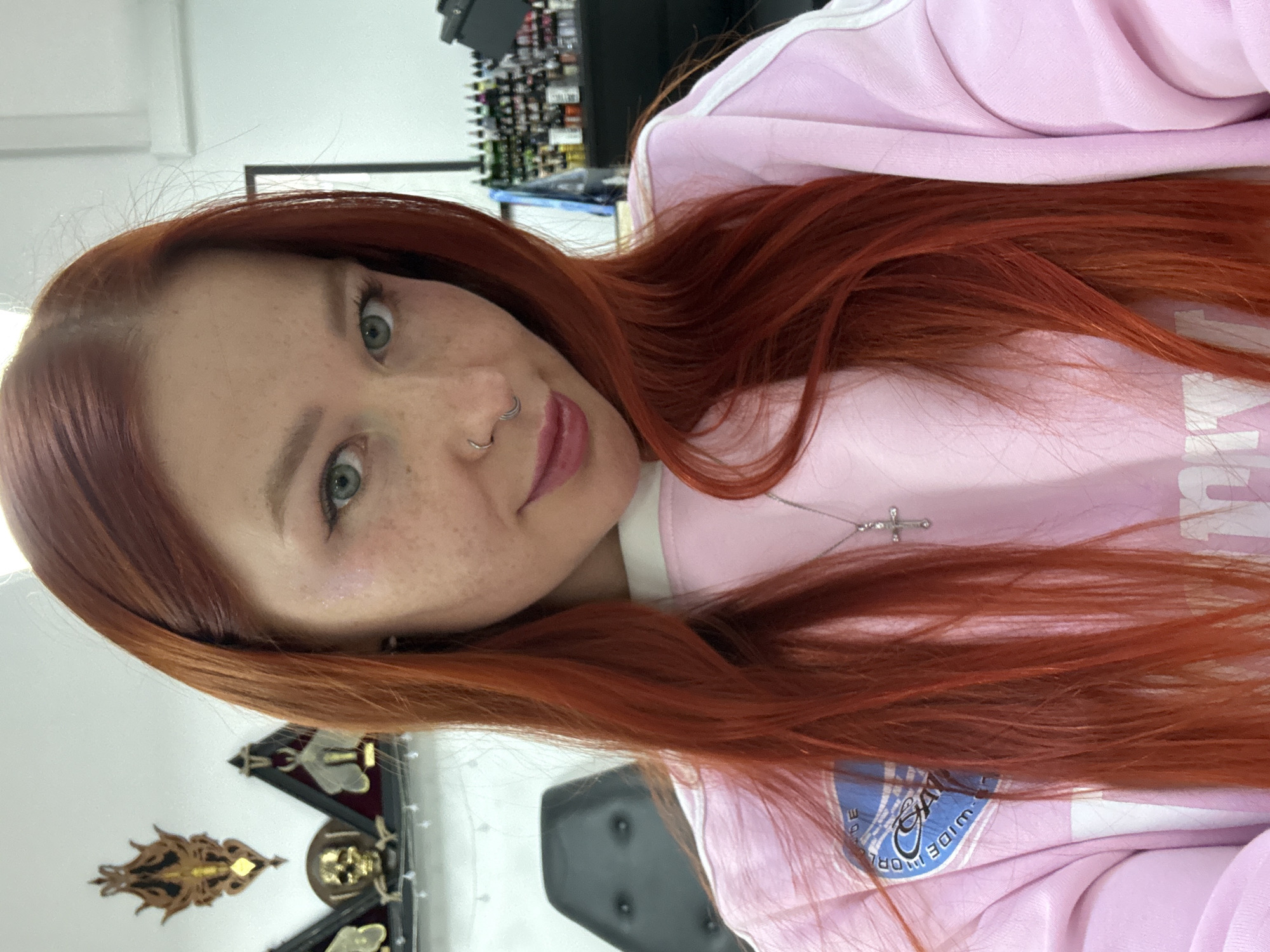 A young woman with long, vibrant red hair and light eyes, possibly green or blue, is captured in a close-up indoor portrait. She has visible freckles across her nose and cheeks. Her nose features a double-hoop piercing on one nostril, and she appears to have another piercing above her upper lip. She is wearing a light pink hooded sweatshirt with white trim, and a thin silver chain necklace with a small cross pendant hangs around her neck.
The woman looks directly at the camera with a calm, soft expression, her lips slightly parted. Her long red hair cascades around her shoulders.
The background, slightly out of focus, suggests an indoor commercial setting, possibly a salon or studio. To the left, a shelf or rack filled with numerous small bottles, potentially beauty products like nail polish or hair dye, is visible. On the far left, behind her shoulder, decorative items are mounted on a wall or shelf, including what appears to be a golden skull and other intricate, metallic designs, some resembling insects. A dark chair or piece of furniture is also partially visible on the right side of the frame.
On her pink sweatshirt, a partial logo or emblem is visible, featuring blue and white colors on the pink fabric. The word "WIDE" and part of another word, possibly "WORLD," are discernible within this design. The location context for this image is Rostov-on-Don, Russia.