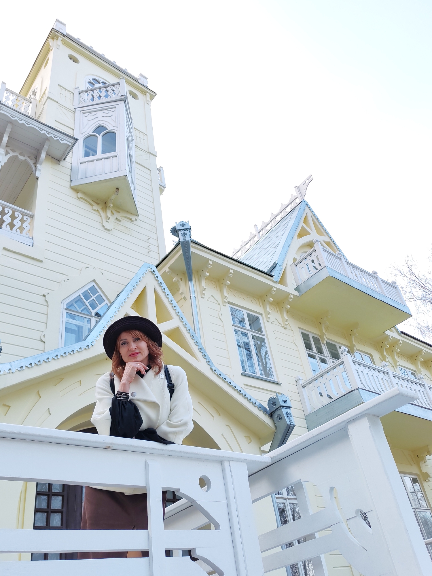 A medium shot presents a red-haired woman wearing a black hat and a light-colored coat, leaning on a white railing. The background showcases a large, elaborate, light yellow building with intricate architectural details. The building has multiple stories, including windows and balconies, with blue-grey accents on the roof and trim. The image was taken in Sarapul, Russia.