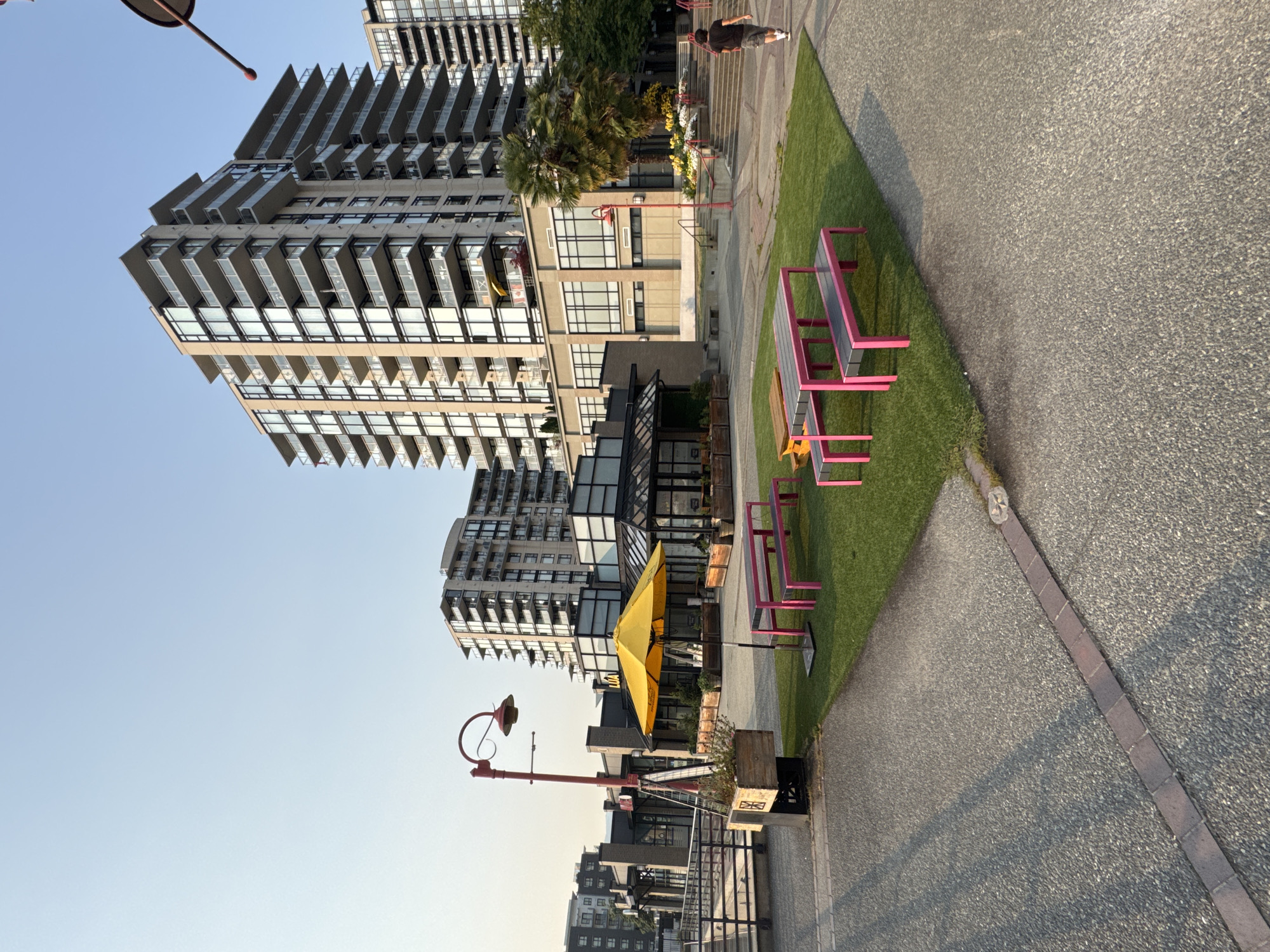A sunlit, outdoor seating area at Lonsdale Quay, North Vancouver, Canada, features modern apartment buildings and a waterfront walkway. Several bright pink picnic tables with dark seating are arranged on a patch of artificial turf. A large, yellow umbrella stands beside one of the tables, casting a shadow. In the background, a person in a dark t-shirt and shorts walks away from the camera along the paved walkway, towards a set of stairs. The Lonsdale Quay Market building, with its large glass windows and exposed beams, is visible to the left of the seating area, and a red lamppost with an old-fashioned lamp stands nearby. The sky is clear and blue, with a slight haze near the horizon.