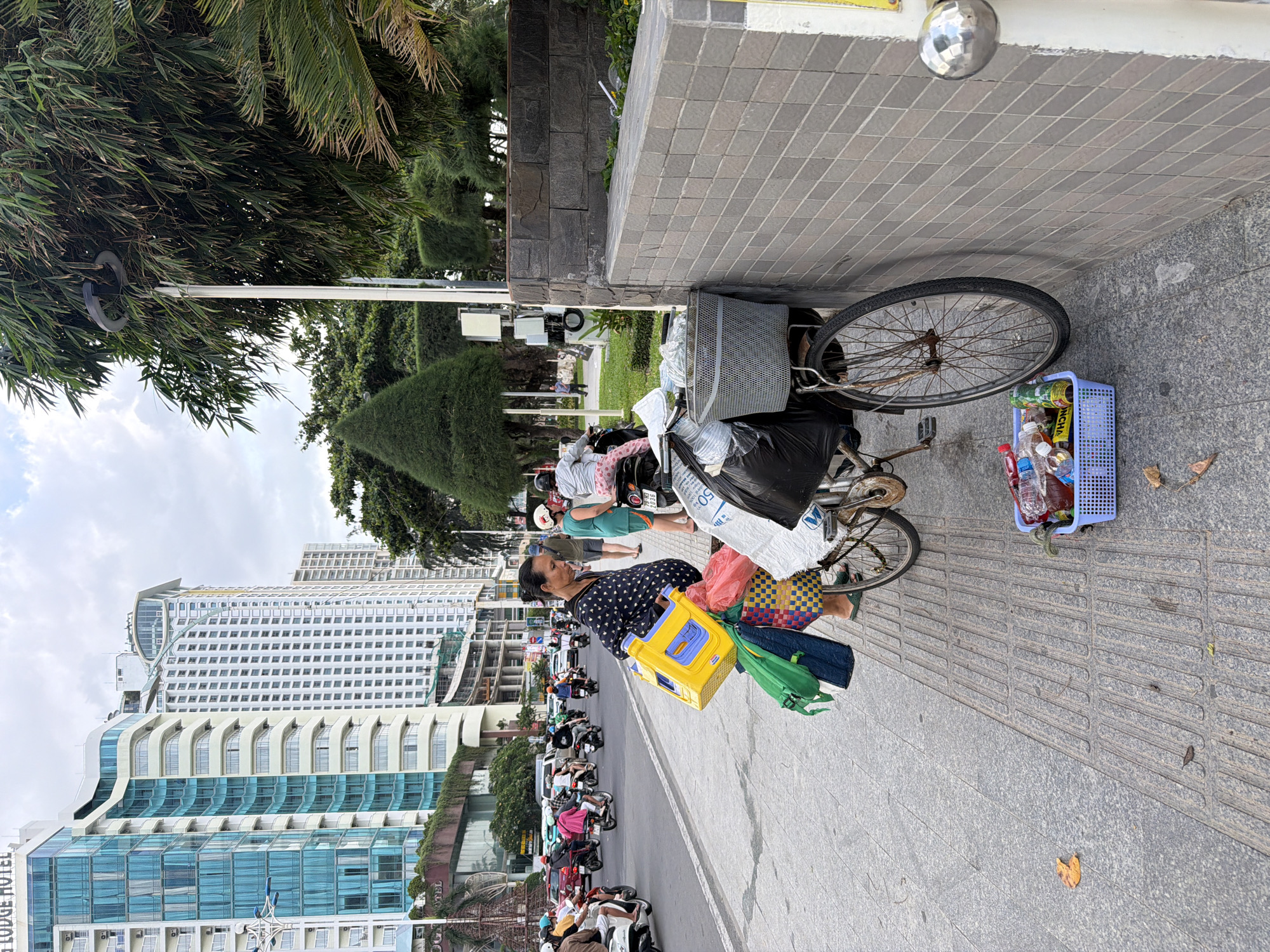 This image depicts a bustling street scene in Nha Trang Ward, Vietnam, characterized by a mix of urban architecture, tropical greenery, and street activity. In the foreground, a woman stands on a wide, tiled sidewalk. She wears a dark blue shirt with white polka dots, dark pants, and flip-flops, holding a bright yellow plastic box and a green fabric bag. She appears to be focused on something to her left.
Beside the woman, a bicycle is parked, heavily laden with various items. Its front basket contains plastic bottles and other goods, while a large black trash bag and several fabric bags, including a white one with "50" partially visible, are secured to the back. A blue plastic basket, with "CHÂN" visible on its side, rests on the ground next to the bicycle, also filled with plastic bottles. The sidewalk features both plain grey tiles and textured tactile paving.
The background reveals a lively street lined with numerous motorbikes, some parked and others in motion, with riders wearing helmets. Tall, modern buildings dominate the skyline, including a multi-story structure where "LODGE HOTEL" is partially visible. Lush green trees, including palm-like foliage and trimmed bushes, punctuate the urban landscape. The sky is bright with scattered clouds, indicating daytime. The overall scene suggests a commercial or mixed-use area, with the woman likely engaged in street vending or transportation.
