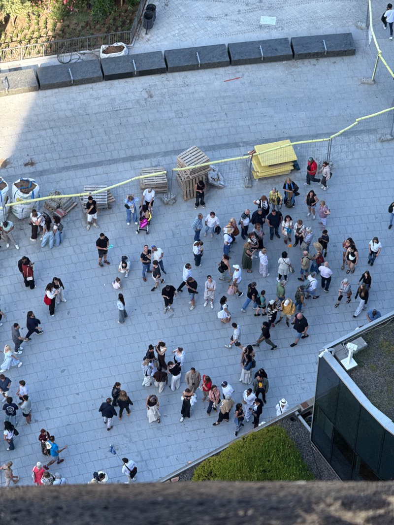 A high-angle, top-down view captures an outdoor urban plaza bustling with numerous people. The scene appears to be a public gathering or a busy thoroughfare, possibly late afternoon or early evening given the long shadows cast by individuals.
The ground is paved with light-colored rectangular pavers. Many people are visible, some standing in small groups seemingly conversing or observing, while others are walking across the plaza. They represent a variety of ages and are dressed in casual attire. In the foreground, several groups are clustered together, and a few individuals are seen walking towards the left and bottom right edges of the frame. One person is pushing a baby stroller.
Throughout the mid-ground and background, there are various objects suggesting an area that might be undergoing construction or temporarily cordoned off. These include stacks of wooden pallets, yellow plastic or metal barriers, large white bags likely containing sand or other construction materials, and a section of yellow caution tape running horizontally across the mid-section of the image. Further back, along the top edge, several large, dark, rectangular concrete blocks are arranged in a line, possibly acting as temporary street barriers or planters.
To the top left, a patch of green foliage is visible behind a fence, suggesting a park or garden area. Another section of green shrubbery and the dark windows of a building are seen along the bottom right edge. The overall atmosphere appears casual and lively. No discernible text is visible on any signs or objects within the image.