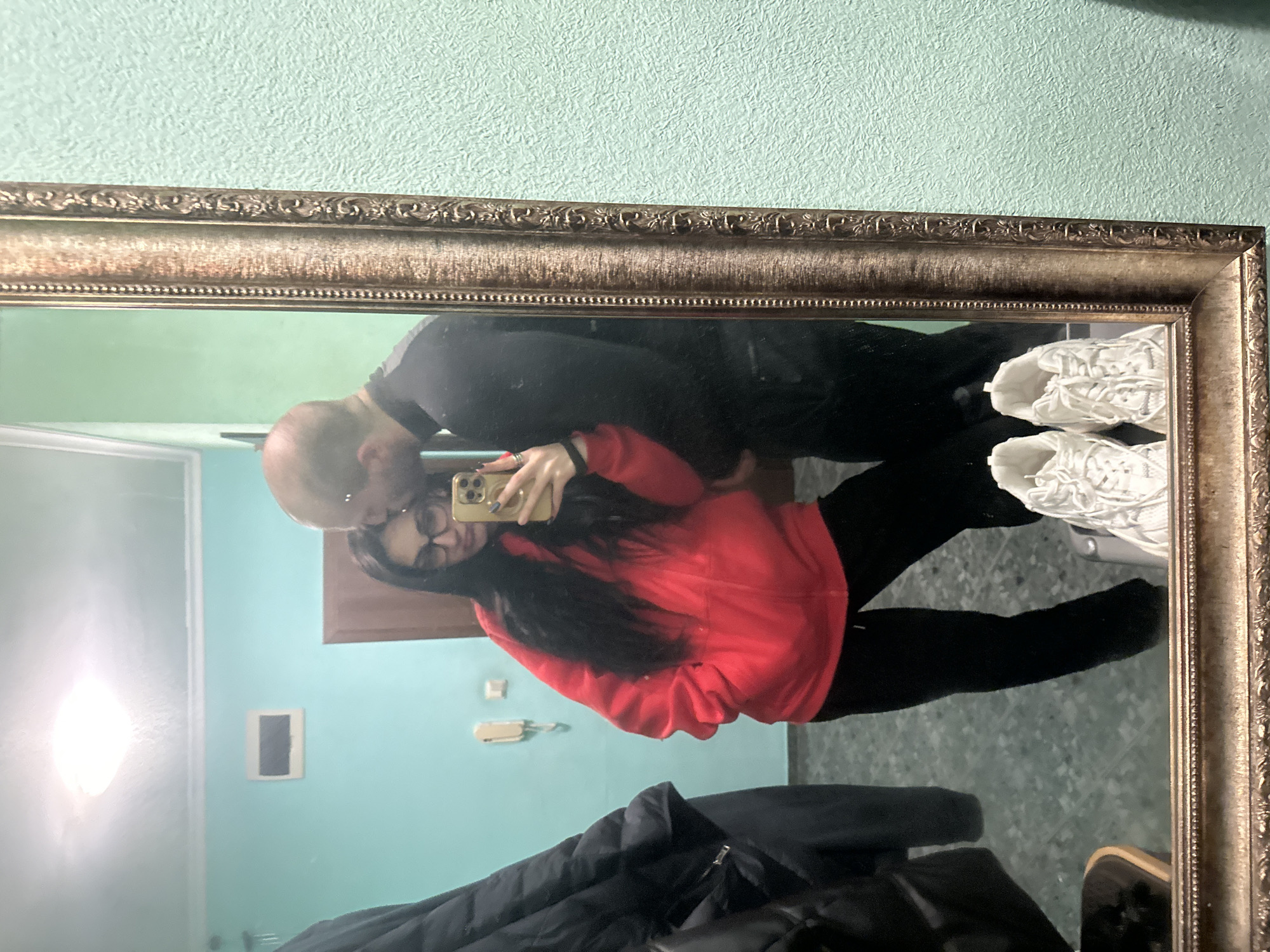 A reflection in a framed mirror shows a man and a woman. The man is bald and wearing dark clothing. The woman has long dark hair, glasses, and is wearing a red hooded sweatshirt and black leggings, she is holding a phone. The background, visible in the mirror, features light blue walls, a door, a dark framed print, and an item that looks like a landline phone mounted to the wall. A pair of white sneakers are on the floor, and a dark-colored jacket is draped.
It is not possible to identify the exact location as Burgas, Bulgaria from the image alone.