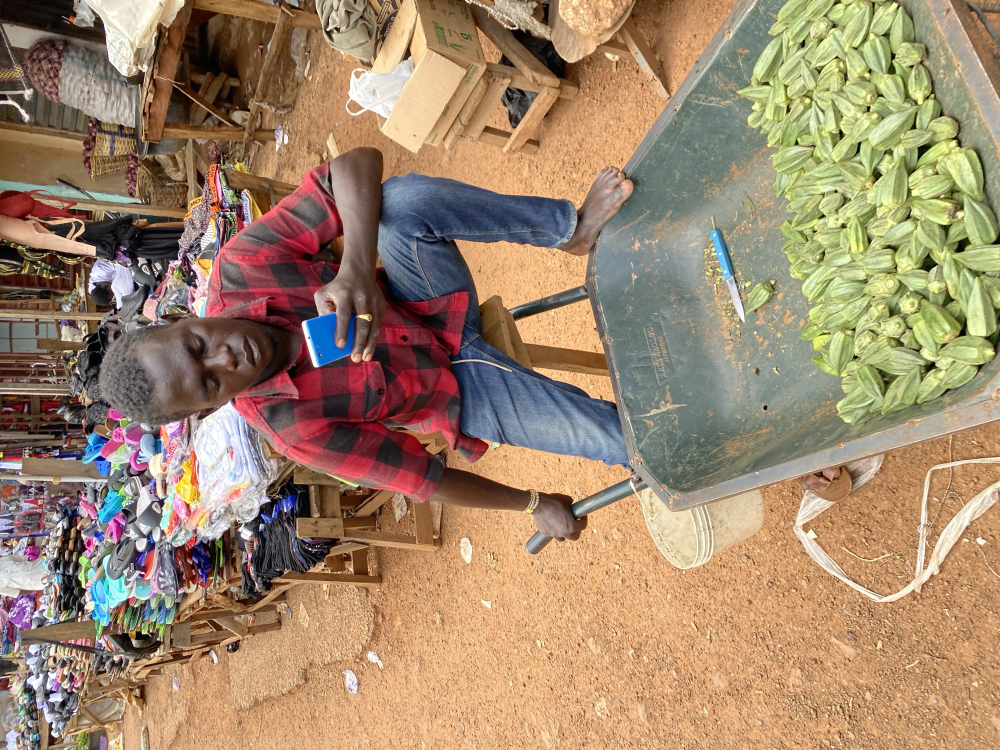 This is a high-angle, full-shot photograph of a man in a market in Bukuru, Nigeria. The man, who appears to be in his late 20s or early 30s, is wearing a red and black checkered shirt and blue jeans. He is holding a blue smartphone in his right hand and a small blue-bladed knife in his left hand, which he is using to prepare okra in a large green wheelbarrow filled with the vegetable. The wheelbarrow is positioned to the right of the frame, and its contents of okra are clearly visible. The man's face is partially obscured, but he appears focused on his task.
The market setting is evident, with various goods displayed in the background, including racks of colorful sandals, clothing, and baskets of produce. The ground is covered in reddish-brown dirt, typical of a dry outdoor market. Several wooden structures and some discarded plastic bags are also visible. The overall impression is of a bustling, vibrant market scene on what appears to be a clear day. No visible text is present in the image. The man's posture and the presence of the smartphone suggest he might be multitasking, possibly communicating or documenting his work while tending to the okra.