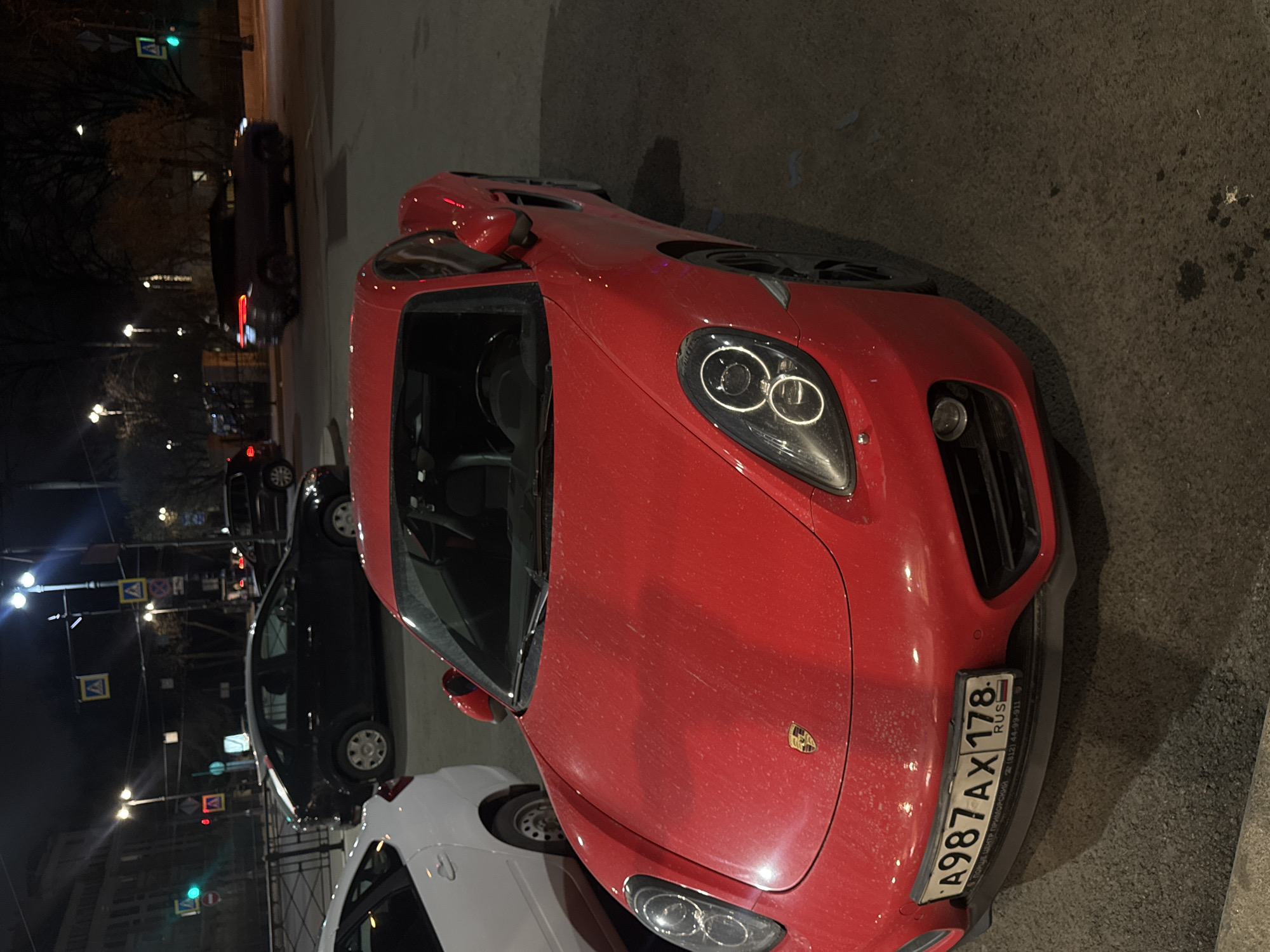 The image captures a street scene at night in what appears to be Saint Petersburg, Russia. Dominating the foreground is a red Porsche sports car, parked on the right side of the frame. The vehicle's front is visible, including its distinct headlights and the Russian license plate A987AX178. The car seems slightly dusty.
Adjacent to the Porsche and slightly behind it, there's a white car. Further in the background, partially obscured by the white car, is a dark-colored sedan. Streetlights illuminate the road, and traffic signs are visible on poles along the sidewalk on the left side of the image. The presence of bare trees suggests the photo was taken in late autumn or winter. The buildings and cityscape in the background provide a sense of urban location.