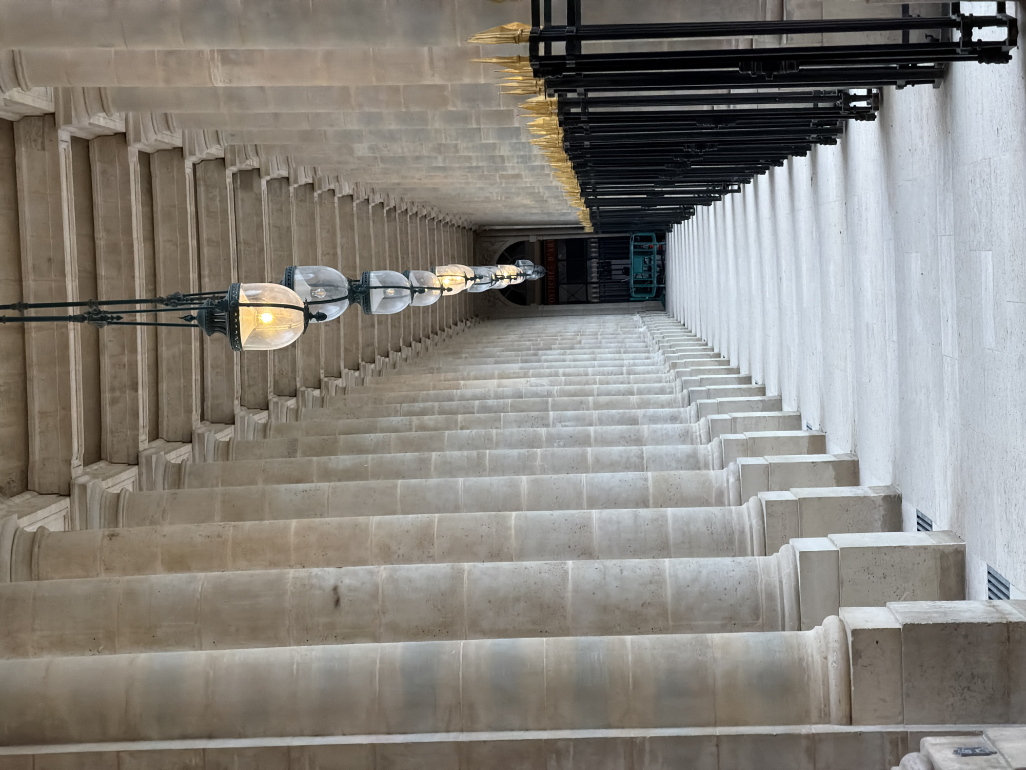 The image shows an upward view of a seemingly endless staircase within a historic building. The architecture is characterized by rows of stacked, off-white stone structures, possibly pillars, which create a tunnel-like effect. On the left, a series of globe-shaped lamps with metal frames hang along the corridor. A black metal barrier runs along the right side of the frame. In the distance, a teal maintenance lift is visible. The location is likely within the Louvre Museum in Paris, France.