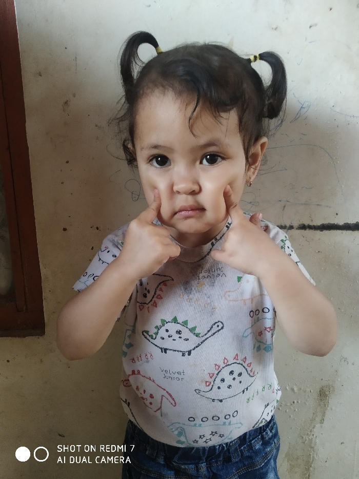 A toddler with pigtails and yellow hair ties is centered in the image. She is wearing a white t-shirt with dinosaur graphics and blue jeans. The child is making a face, pushing her cheeks inward with her index fingers. The background is a plain wall with some scuffs and a dark window frame on the left. The words "SHOT ON REDMI 7 AI DUAL CAMERA" are visible in the bottom left corner.