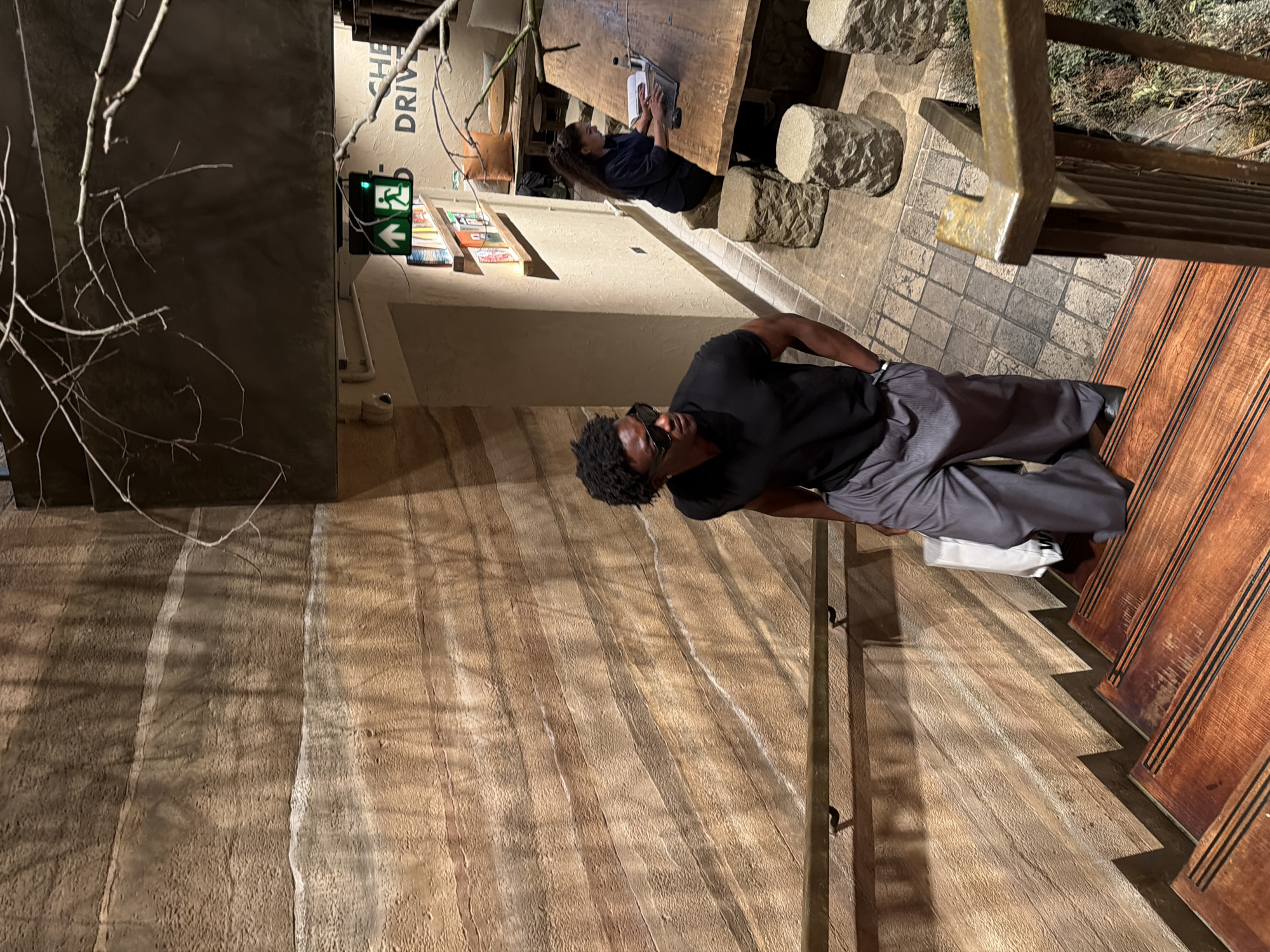 Here is a description of the image:
The photo shows a man standing on a set of wooden stairs, likely outdoors. He is wearing sunglasses, a black t-shirt, and gray pants. Another person is visible in the background sitting at a table, using a laptop. The setting appears to be a cafe or restaurant, identifiable by the wooden table, stone seating, and a sign with the word "DRIVE" partially visible. There's an exit sign with an arrow pointing to the left, suggesting the location is in the United Kingdom. A branch of a tree is visible in the foreground. The lighting is dim and mostly comes from overhead sources.