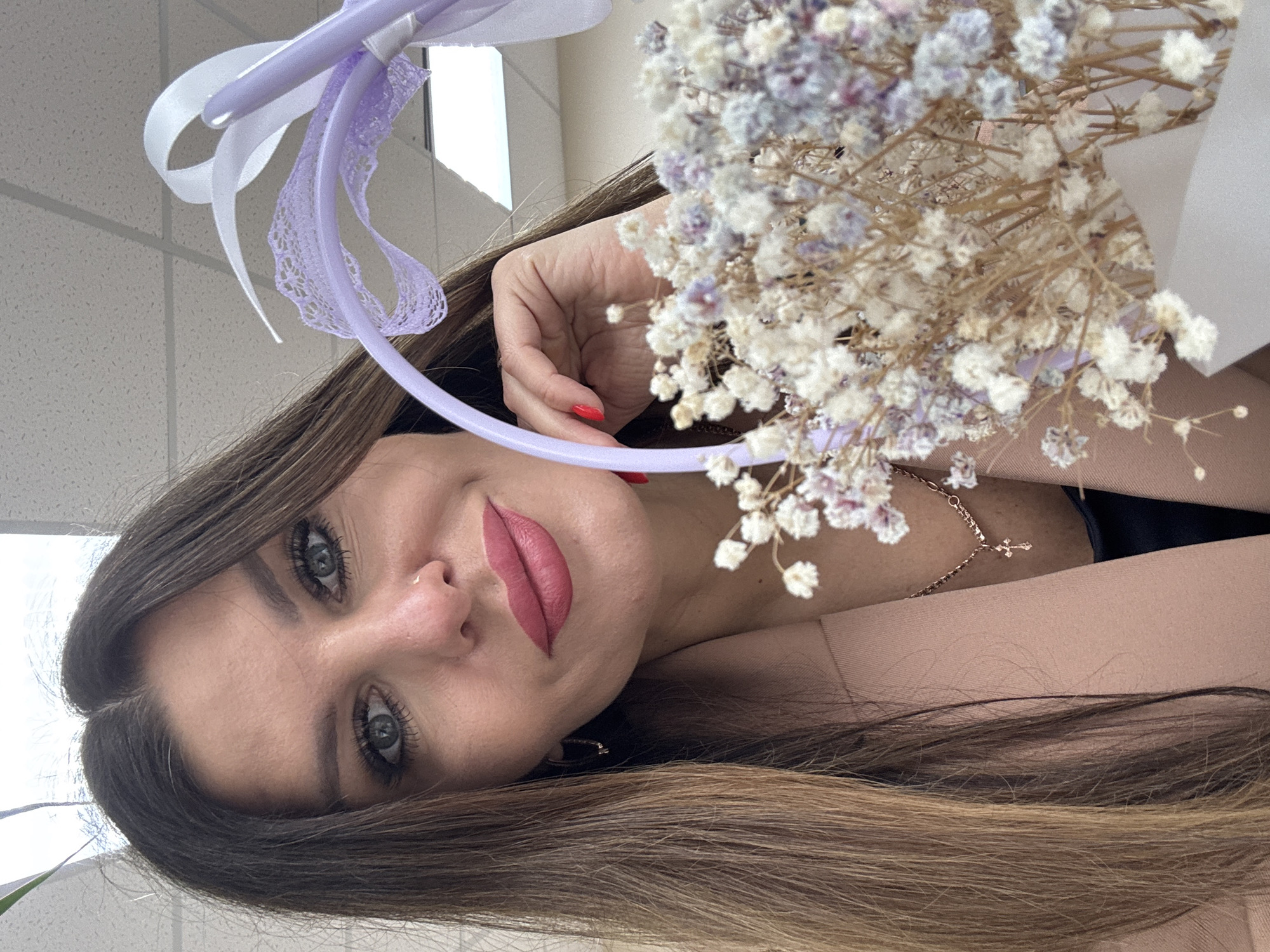 The image shows a woman holding a bouquet of dried baby's breath flowers. She is wearing lipstick and has long brown hair. A purple hoop with white and purple ribbons is visible behind the flowers. A gold necklace with a cross pendant is around her neck. The background appears to be an indoor setting with a white tiled ceiling. The location is Chaikovsky, Russia.