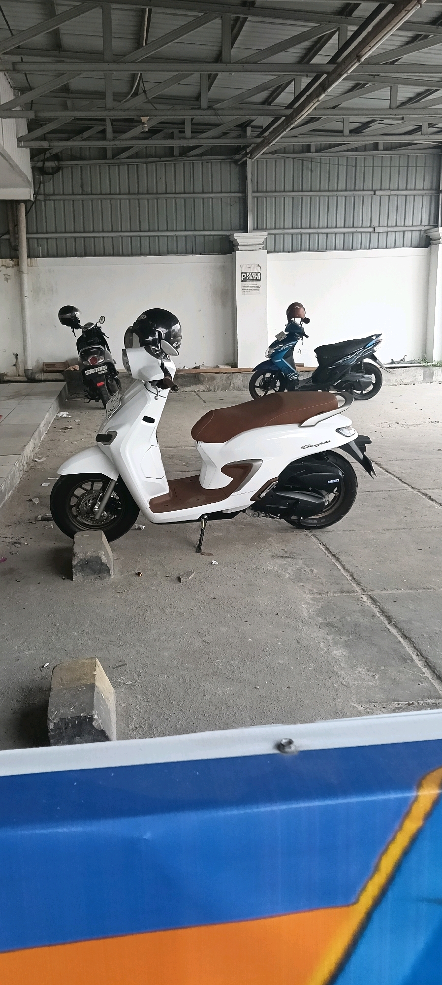 The image depicts a covered parking area in Kuala Kapuas, Indonesia, featuring several parked scooters. In the foreground, a white scooter with a brown seat is prominently displayed, with a black helmet resting on its handlebars. To the left, another scooter, dark in color, is parked with a helmet on its seat. Further back, a blue scooter is visible, also with a helmet on its seat. The parking area is roofed with a corrugated metal structure supported by metal beams. The walls are white, and a sign with "PARKIR GRATIS" (free parking) is affixed to the wall. The ground is paved with concrete, and there are some debris scattered around. The scene is observed from a slightly elevated, eye-level perspective.