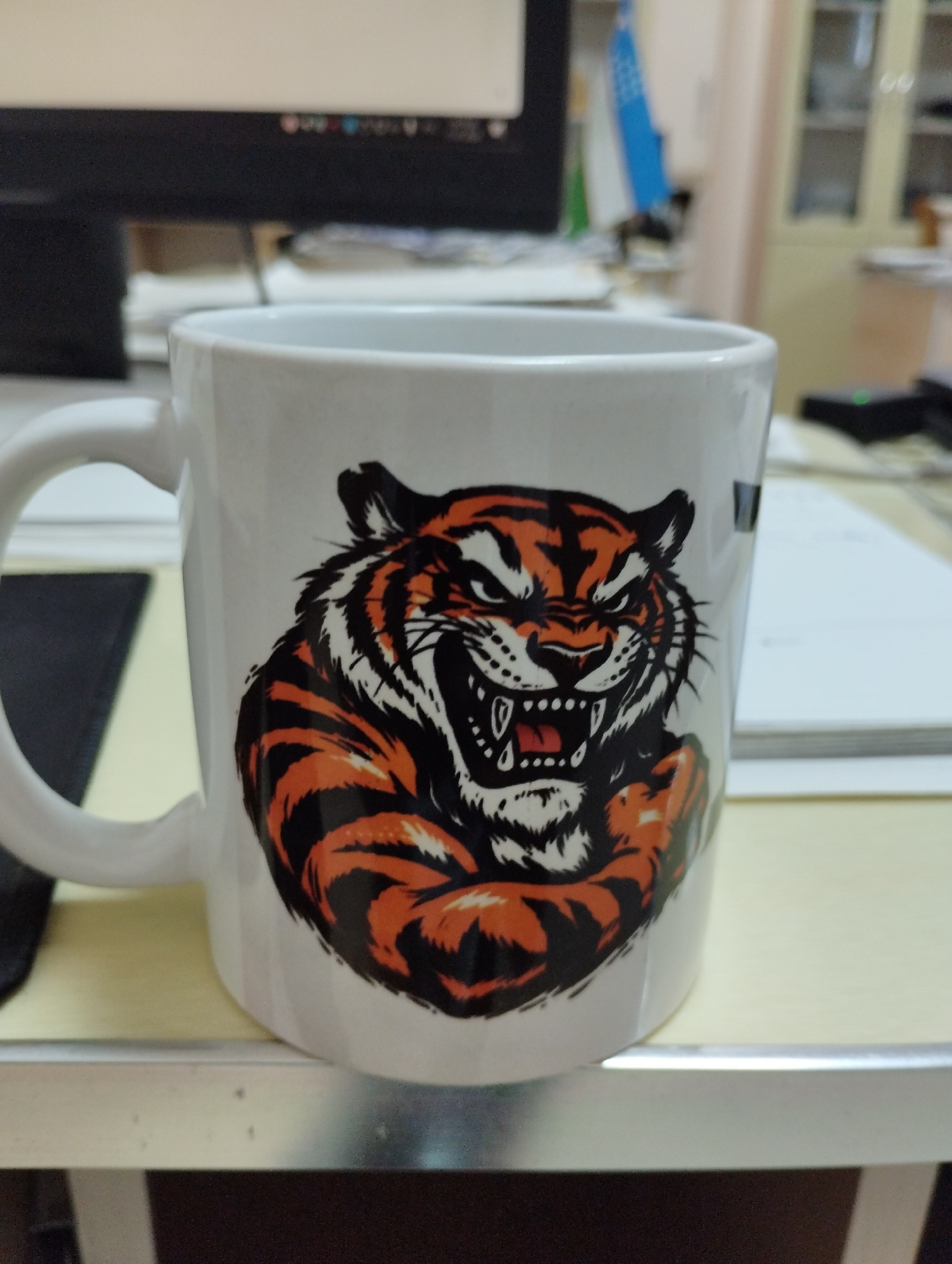 A white mug with a roaring tiger graphic is the primary subject, positioned centrally in the frame. The mug features the tiger's head and chest, depicted with sharp teeth, an open mouth, and striking orange and black stripes. The background is blurred, suggesting an indoor setting, possibly an office or workspace. Visible elements in the background include a computer monitor, papers, and office furniture, hinting at a professional environment. The overall lighting suggests ambient indoor illumination. The image is taken from a slightly elevated perspective, focusing on the mug. There is no information about the city of Urgench, Uzbekistan.
