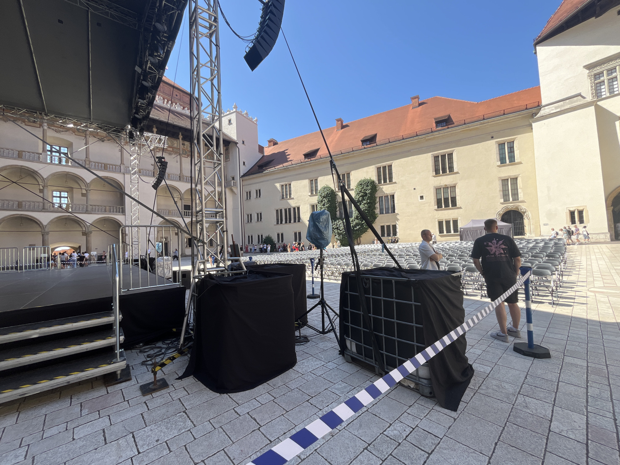 The image depicts an outdoor setup for an event, likely a concert or performance, in a courtyard with historic architecture. The scene is set in Krakow, Poland, specifically within the Wawel Castle courtyard.
In the foreground, a stage structure is partially visible on the left, with steps leading up to it. Several large black fabric-covered objects, possibly speakers or equipment, are positioned near the stage. A blue and white striped barrier tape is laid out across the paved ground, creating a pathway or demarcation. Two men are standing near the equipment, one in a black t-shirt and shorts, the other in a white t-shirt.
The background reveals rows of empty chairs set up on the cobblestone courtyard, indicating a seating area for an audience. The architecture surrounding the courtyard features buildings with arched windows and red-tiled roofs, typical of historic European structures. The sky is clear and blue, suggesting a sunny day. People are visible in the distance, either setting up or mingling before the event. The overall scene suggests preparation for a cultural event in a historically significant location.