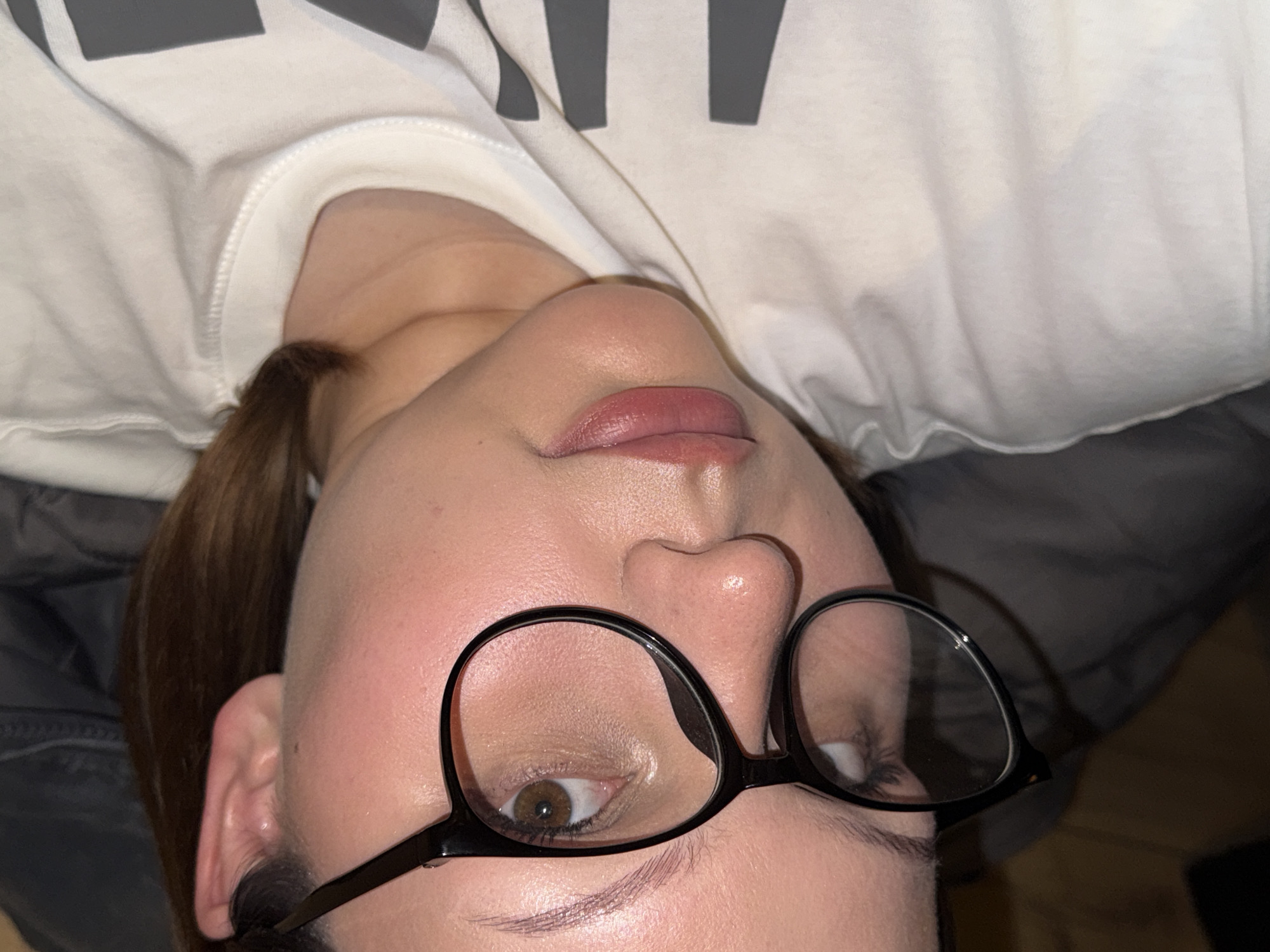 A close-up, upside-down, high-angle shot shows a woman lying down wearing black-rimmed glasses, a white shirt with gray lettering, and resting on what appears to be gray fabric. The location of the image is not provided and therefore cannot be deduced.
