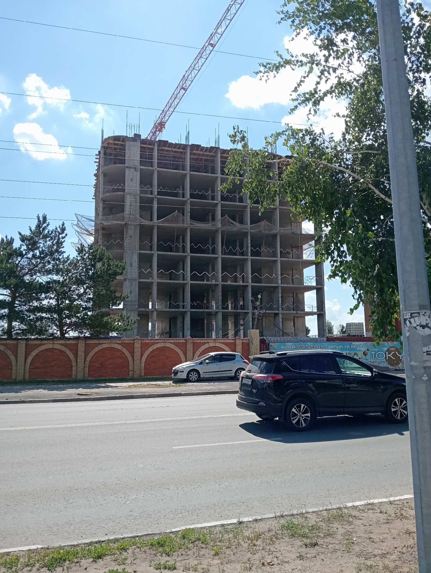A construction site with a partially built concrete building and a large red crane is visible under a clear blue sky with scattered clouds. A dark SUV and a silver sedan are parked on the side of a paved road in front of a brick fence. Trees line the street, and a tall grey pole stands to the right of the frame. A poster with text in an unfamiliar script is attached to the pole.