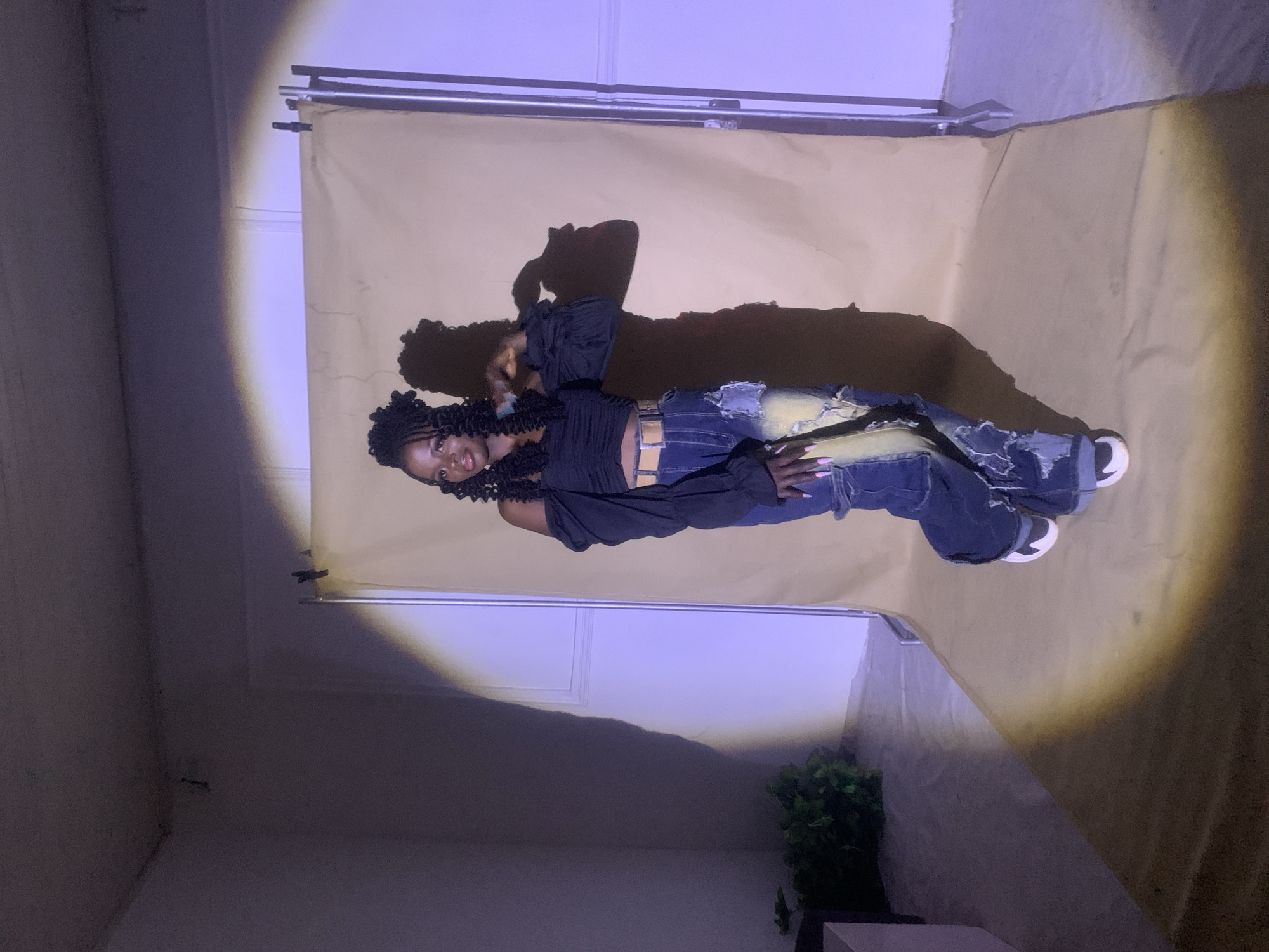 A full-length portrait of a Black woman with long, braided hair stands in front of a beige backdrop in a room with white walls. She is wearing a black, off-the-shoulder top, distressed denim jeans, a brown belt, and white sneakers with black accents. A shadow is cast on the backdrop behind her. A plant is visible in the corner of the room. The lighting appears to be from a single source above, creating a strong spotlight effect. No definitive evidence places this photo in Jos, Nigeria, and this cannot be confirmed.
