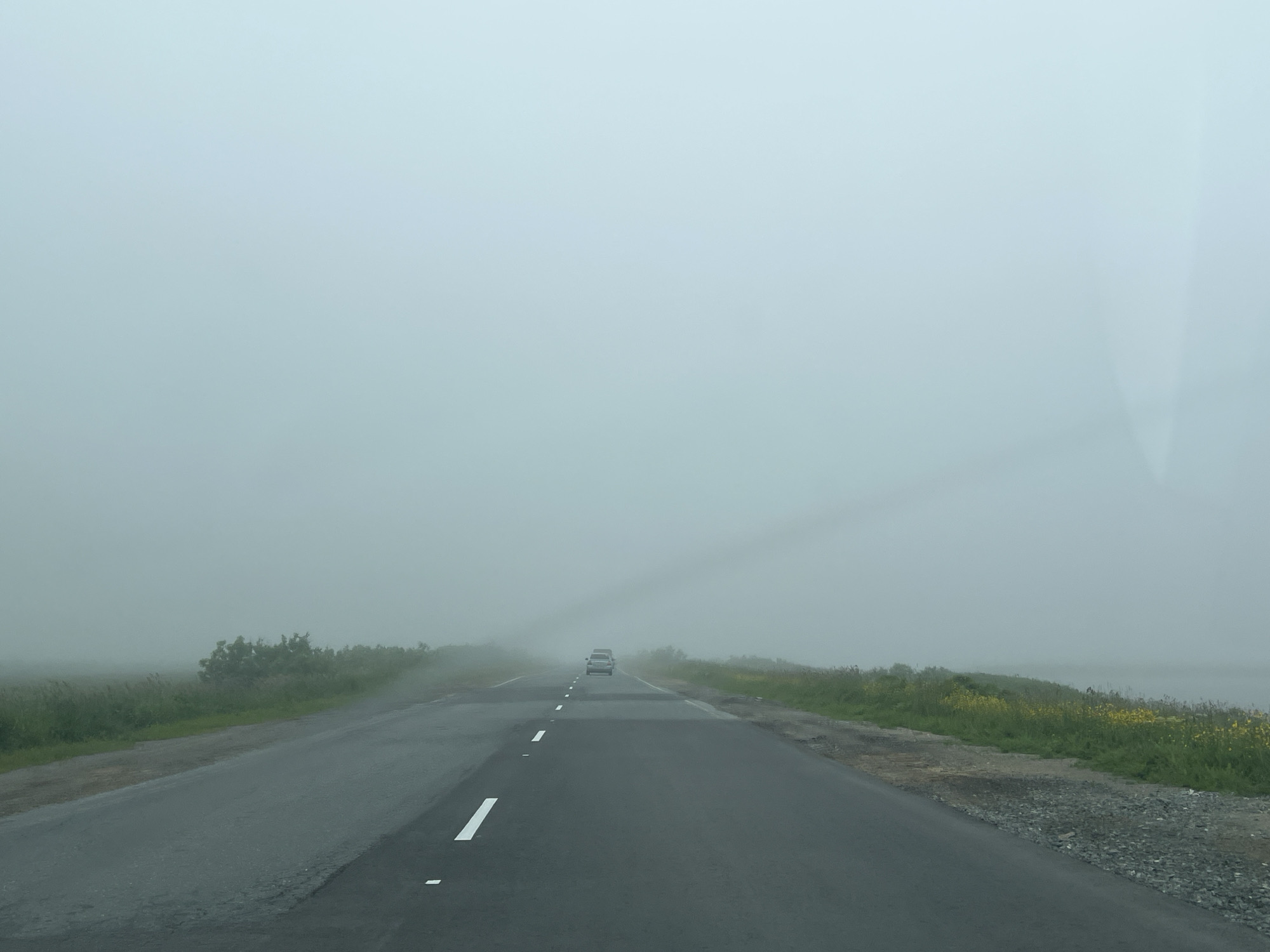 The image depicts a road in a foggy environment. The road is paved and has white lane markings. On either side of the road, there is vegetation, including grass and some bushes or trees. In the distance, a car is visible traveling on the road, partially obscured by the fog. The sky is overcast and appears very hazy, contributing to the overall low visibility. There are no people visible in the image. The scene suggests a rural or semi-rural setting with limited infrastructure. The specific location in the city of Sovetskoe, Russia, cannot be determined from the visual information alone.