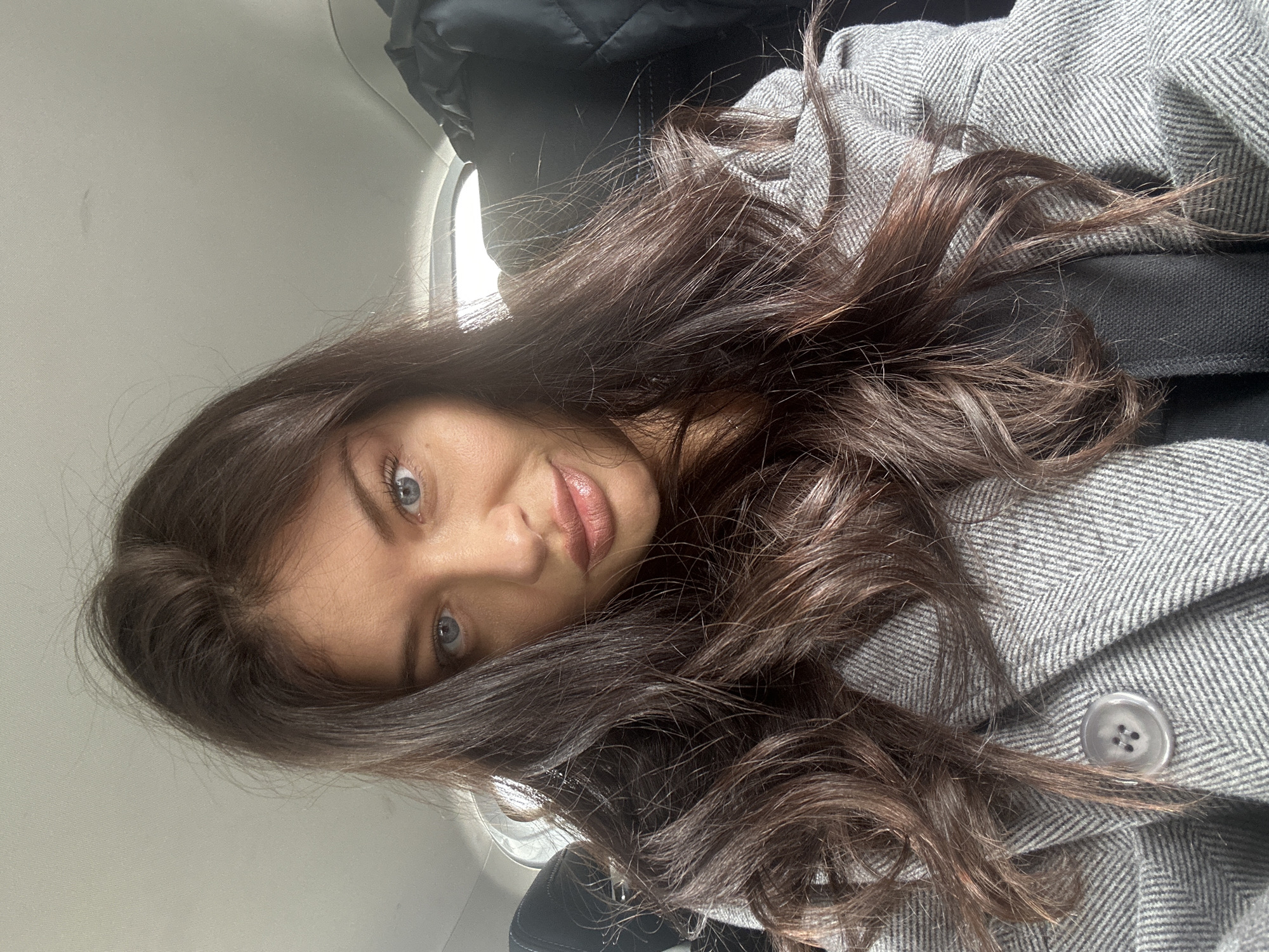 A close-up image shows a young woman with long, wavy dark brown hair and striking blue eyes, looking directly at the camera with a calm expression. She has fair skin and full, light pink lips. She is wearing a grey coat with a herringbone pattern, and a single light grey button with four holes is visible on its front. A dark, possibly quilted, garment or seatbelt strap is visible behind her shoulder.
The setting appears to be inside a vehicle, likely an airplane or train, suggested by the small, bright window visible in the upper right background and the light-colored, plain interior wall/ceiling. The bright light from the window indicates it is daytime.
The image is oriented vertically, with the woman's head positioned towards the right side of the frame, implying she is reclining or lying down while taking the photograph. The scene captures a moment of travel. No text is visible in the image. The geographical context for this scene is Krasnogorsk, Russia.