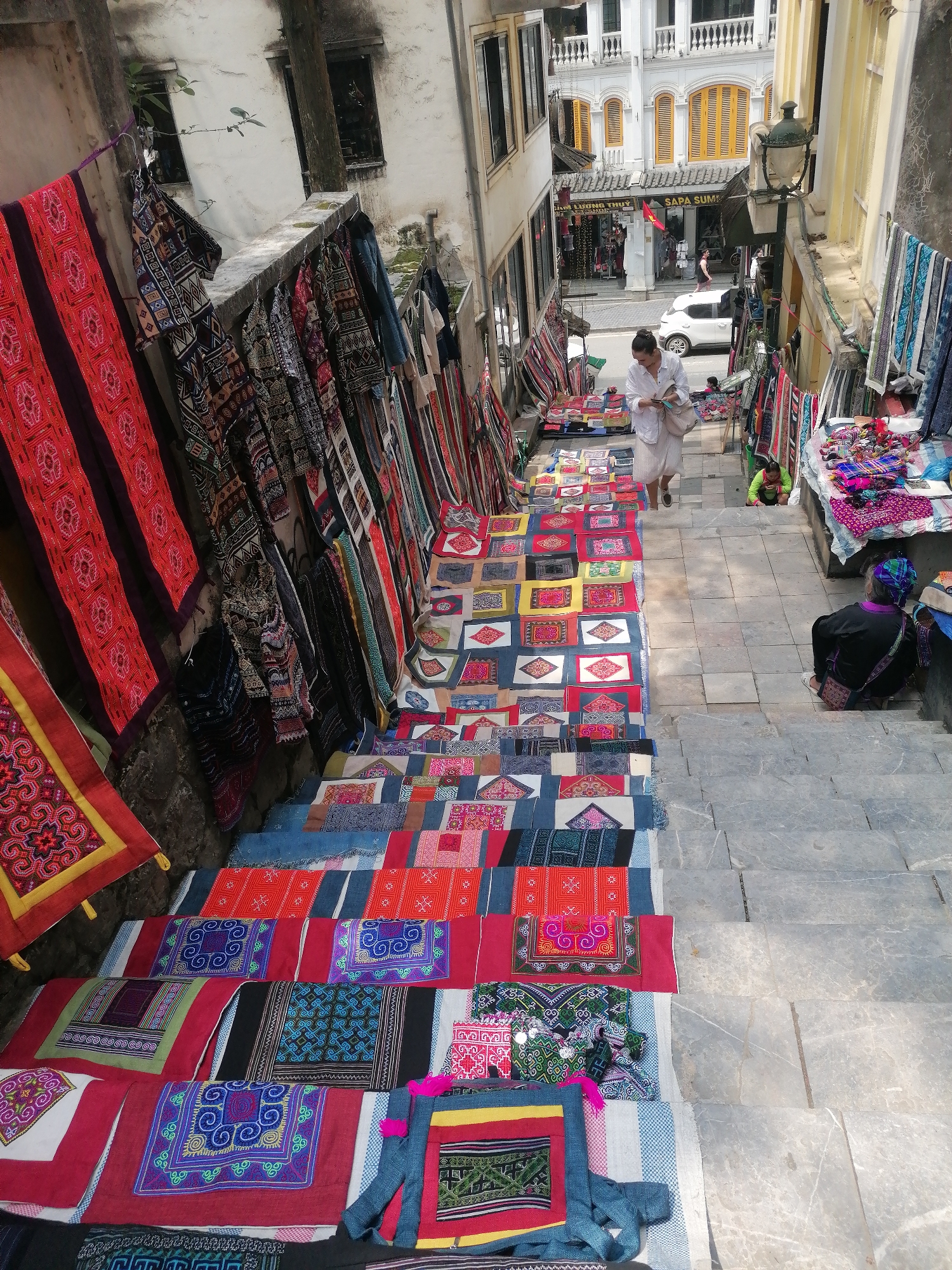 A street market scene in Sa Pa Ward, Vietnam. Numerous colorful textiles are displayed on the steps of a downward-sloping alleyway. More textiles hang from the walls on either side. A woman in white walks down the steps, looking at her phone. An older woman in traditional clothing sits on the steps to the right. Another vendor kneels further up. A white car and several people are visible at the top of the alley. Buildings flank the alley. A lamp post stands to the right.