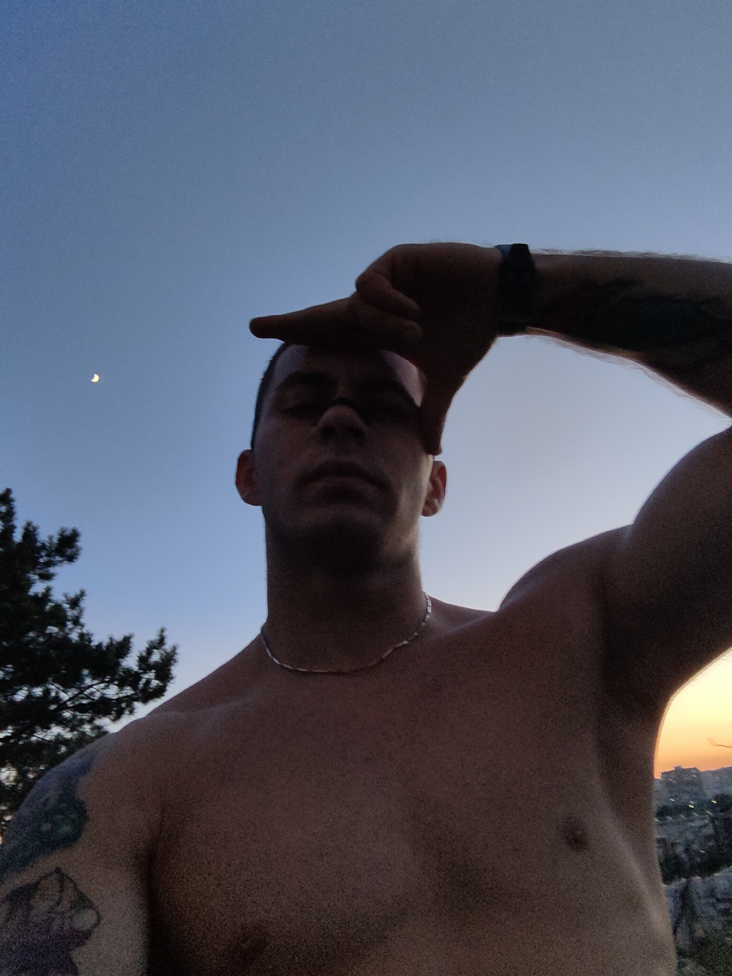 A low-angle, close-up, outdoor, selfie-style shot shows a shirtless man against a twilight sky. The man, with short dark hair and a silver chain necklace, is holding his right hand up to his forehead, framing his face. He has tattoos on his left arm and right arm, and a dark band on his right wrist. The sky is a gradient of pale blue to orange at the horizon. A crescent moon is visible in the upper left quadrant of the sky. To the left, a dark green tree is partially visible. In the background, at the horizon, a city skyline with buildings is illuminated by the setting sun. The man's skin is tanned, and his chest and shoulders appear muscular. The image may have been taken at the Varna Necropolis, a historical site overlooking the city of Varna, Bulgaria, known for its ancient Thracian tombs and panoramic views of the Black Sea and the city.