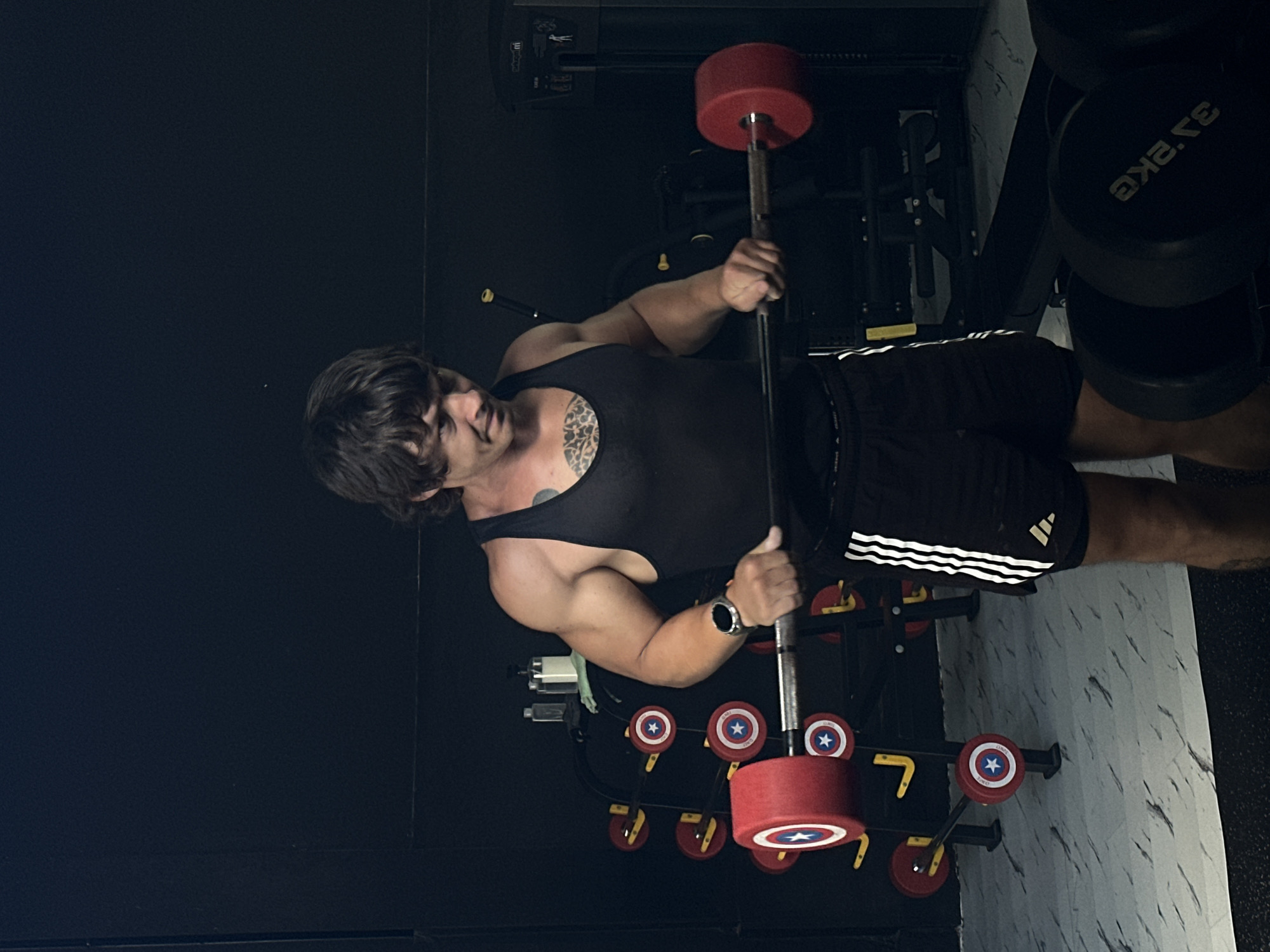 A man is in a gym, holding a barbell loaded with Captain America-themed plates. The man is wearing a black tank top and Adidas shorts, and he has a tattoo on his chest. The gym has a black wall and white marble-patterned flooring. Weight racks with Captain America-themed plates are visible in the background. The photograph was taken in a gym at Tibubeneng, Indonesia.