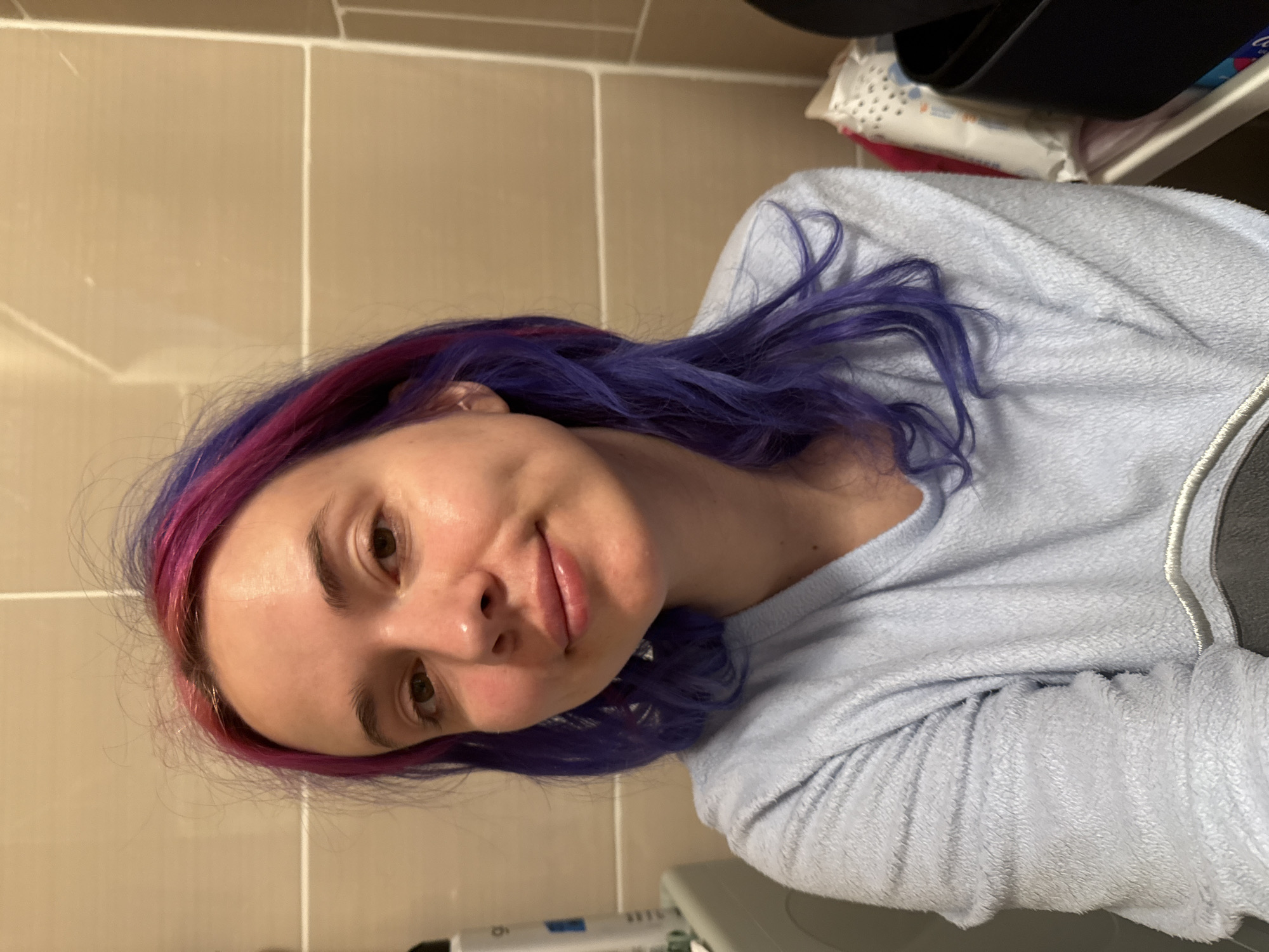 A woman is depicted in a close-up, chest-up portrait, taken indoors, likely in a bathroom setting within the City of Zagreb, Croatia. She has fair skin, dark brown eyes, and a slight, gentle smile. Her hair is vibrantly colored, a mix of bright purple and pink, styled in loose, wavy strands that frame her face. She appears to be wearing no visible makeup, and her skin has a natural, slightly shiny appearance.
She is dressed in a light blue, long-sleeved top made of a soft, fluffy, or fleece-like fabric. A silver-colored trim or embroidery detail is visible along the collar or shoulder seam on the right side of her top.
The background features light beige or tan rectangular tiles with white grout lines, consistent with bathroom wall tiling. In the upper right corner, behind the woman, there's a glimpse of a dark object, possibly a bin or shelf, and beneath it, several white packaged items, resembling toiletries or wipes, are visible. A blurry white object with illegible blue text can be seen in the bottom left, possibly part of a counter or appliance. The lighting appears to be artificial indoor lighting. The woman projects a relaxed and calm demeanor, looking directly at the camera with a soft expression.