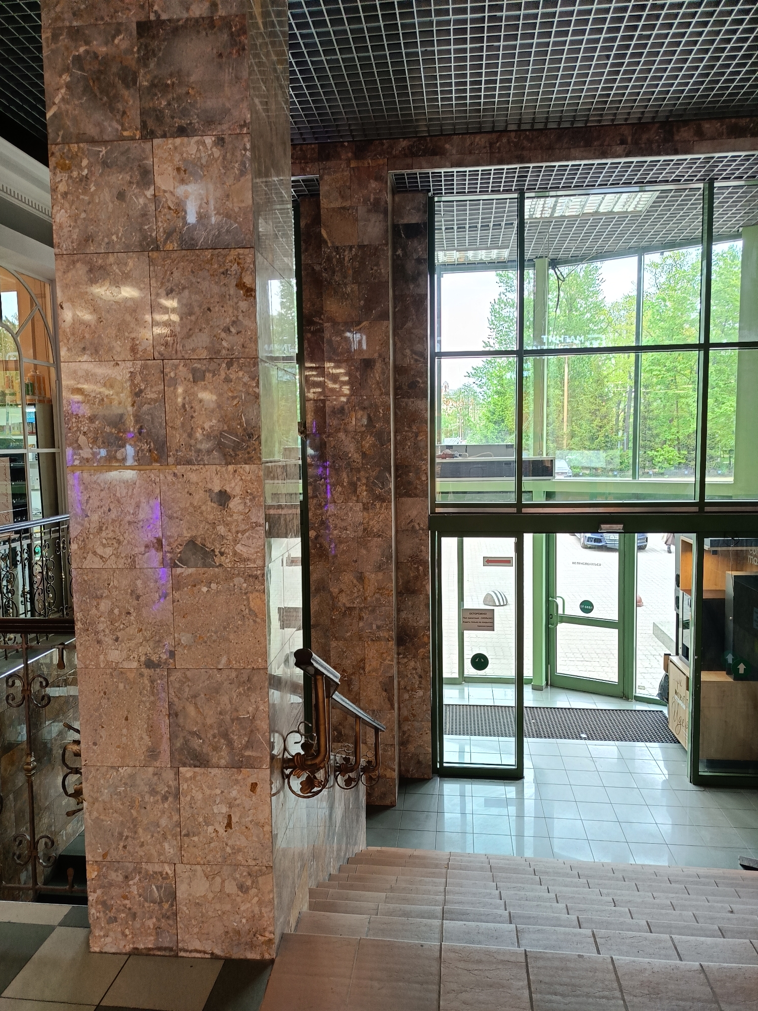 Captured from an elevated position, the image showcases a modern indoor setting, leading to an entrance of a building likely in Saint Petersburg, Russia. The interior features a multi-story structure, apparent from the staircase extending downwards from the foreground. The walls and columns are clad in large, rectangular marble tiles of varying shades of brown and beige, adding a touch of elegance and sophistication to the space.
To the left, an ornate wrought-iron railing suggests a higher level overlooking the main area. The staircase itself is also constructed of marble, blending seamlessly with the walls.
The focal point of the image is the entrance, characterized by large glass windows and doors framed in a vibrant green. Through the glass, one can observe glimpses of the outdoors, including trees and possibly a street with parked cars. Signage is visible on the doors, indicating that they are the main entrance and exit.
The ceiling above is a grid pattern, further accentuating the modern architectural design of the building. Overall, the scene depicts a well-maintained and aesthetically pleasing interior space, designed with a blend of natural materials and modern elements.