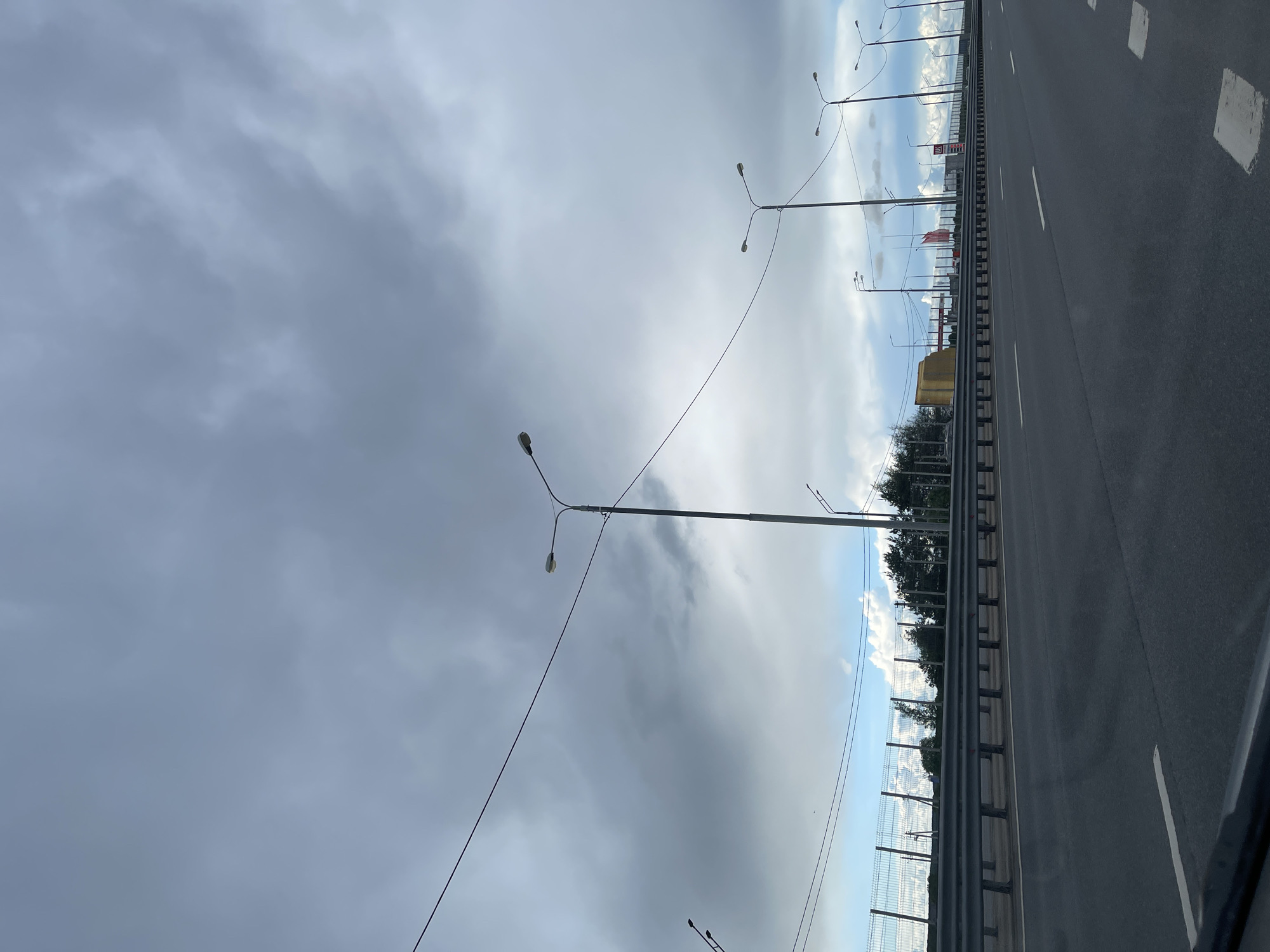 The image shows a view from a highway overpass in Бавыкино, Russia. The highway runs horizontally in the image with multiple lanes. Guardrails line the side of the road, with trees and buildings visible beyond. Overhead streetlights run along the highway. The sky is overcast with grey clouds, suggesting a rainy or cloudy day. There are no people visible.
