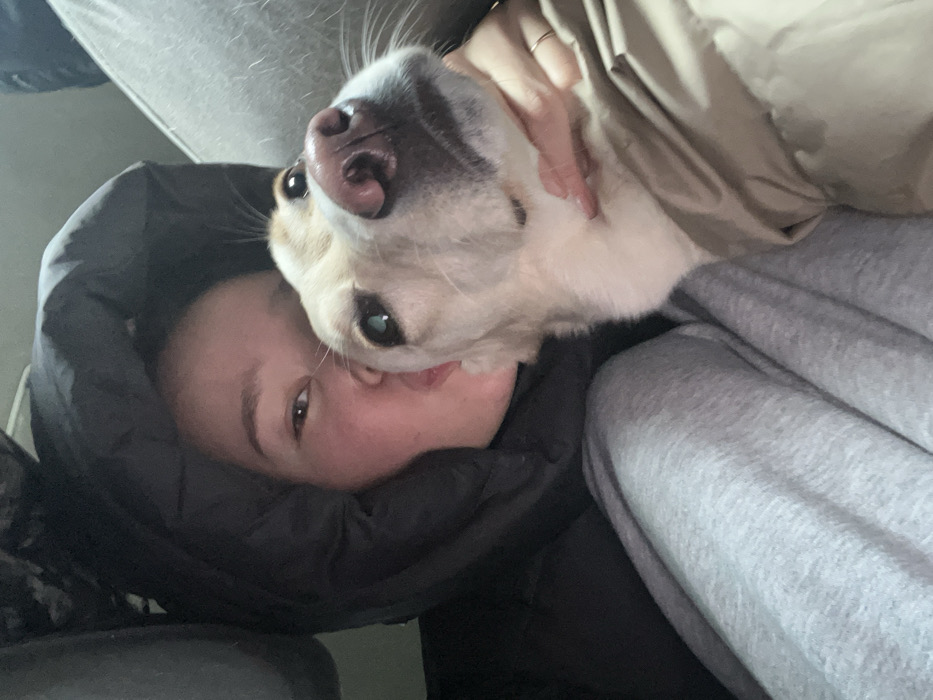 A medium close-up shot captures a person and a dog within an indoor setting at Yemelyanovo Airport, Russia. The human subject, a female, is positioned on the left, wearing a black hooded puffer jacket with the hood obscuring much of her head. Her face, featuring light skin and dark eyes, is visible from the eyes down, directed slightly right of the camera with lips pursed in a kissing gesture. She is also wearing grey sweatpants. Her left hand, adorned with a thin gold ring, supports the neck of a light-colored dog. The dog, appearing cream or light tan, occupies the right side of the frame. It wears a light brown or tan coat, has a dark pinkish nose, and its dark eyes are directed upwards and slightly left. Its head is tilted upwards towards the camera. The background consists of indistinct grey fabric, consistent with seating or interior paneling.