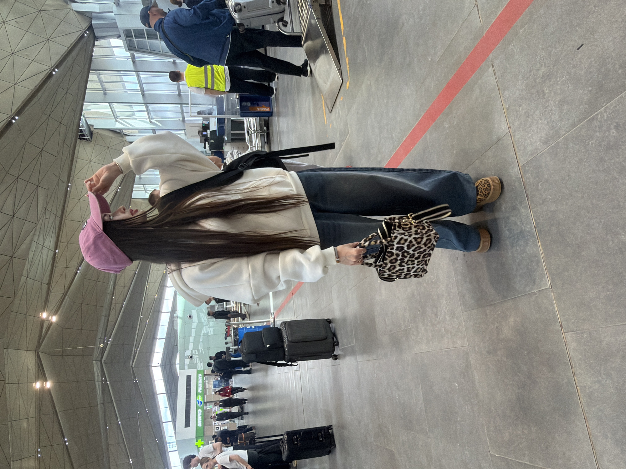 The image, set in what appears to be an airport or large transit hub in Saint Petersburg, Russia, captures a woman in the foreground and a queue of people behind her. The woman, viewed from the side, has long dark hair flowing down her back. She wears a pink baseball cap, a white long-sleeved hoodie, loose blue jeans, and distinctive leopard-print platform shoes. She carries a small leopard-print handbag in her right hand and has a black backpack slung over her left shoulder.
Behind her, a queue of travelers with various pieces of luggage, including black suitcases and backpacks, stretches into the background. Some individuals are visible further down the line, including staff members, one wearing a high-visibility yellow vest. The floor is made of light gray tiled concrete, marked by a prominent red line that extends across the scene. The building's interior features a high, geometrically patterned ceiling and large windows that allow natural light into the spacious area. Overhead lights are also visible embedded in the ceiling structure.
The activity suggests people are waiting in line, likely for check-in, security, or boarding. In the background, blurry green signs can be seen, one of which appears to read "PHARMACY" or "APTEKA" (Russian for Pharmacy). Another blue object in the mid-ground has partially visible text that could be "СУМКА" (SUMKA - bag). The scene depicts a modern, well-lit terminal environment during what appears to be daytime.