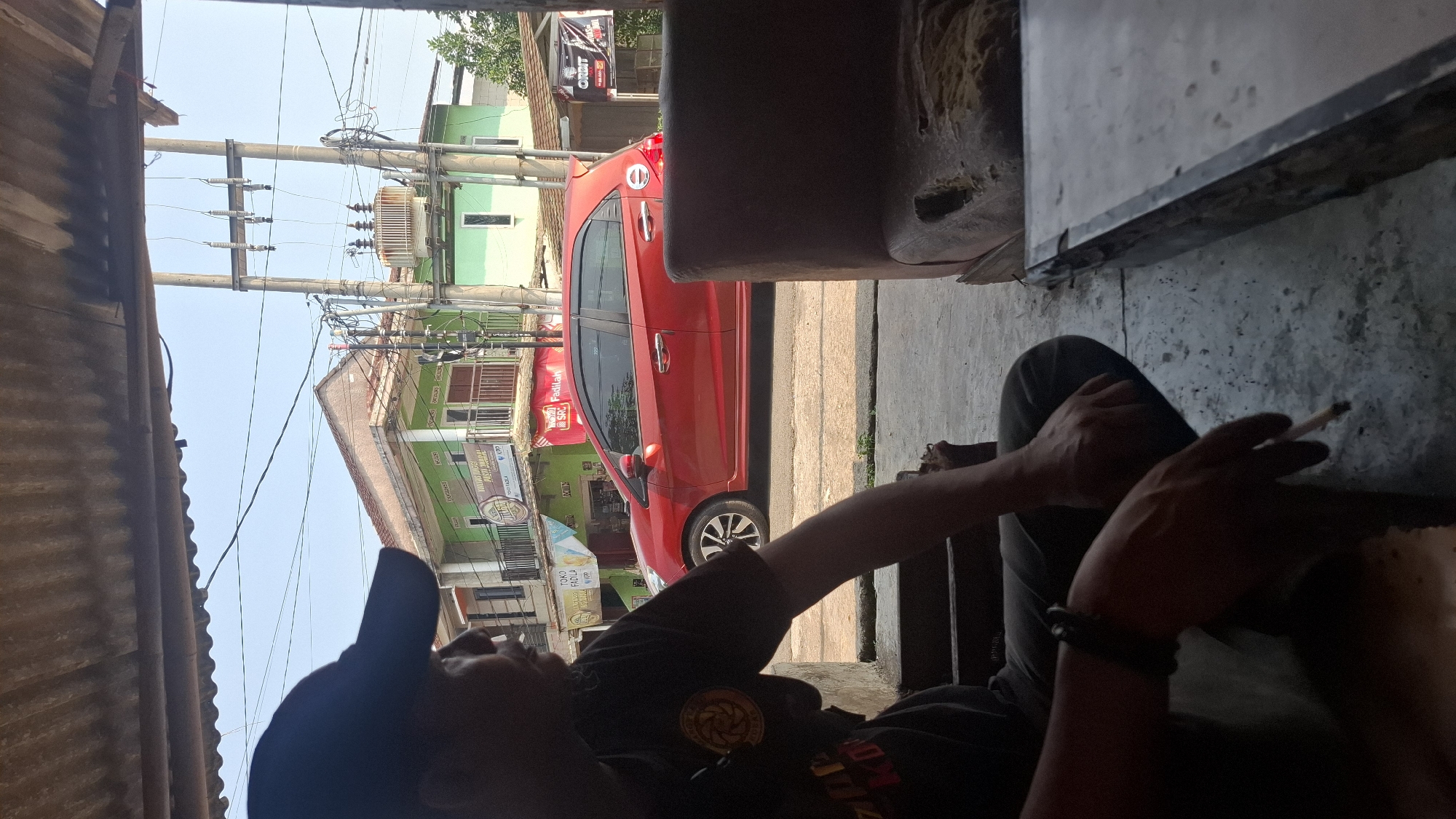 The image shows a man in a dark t-shirt and baseball cap sitting in what appears to be an outdoor seating area with a corrugated metal roof. He is holding a lit cigarette in his right hand, which is resting on his lap. His left arm is bent, and his left hand is resting on the arm of a chair or bench. In the background, a red car is parked on the side of a street. Several buildings and electrical poles are also visible, suggesting an urban or semi-urban environment. Based on the context provided, this scene is located in Cilaku, Indonesia.