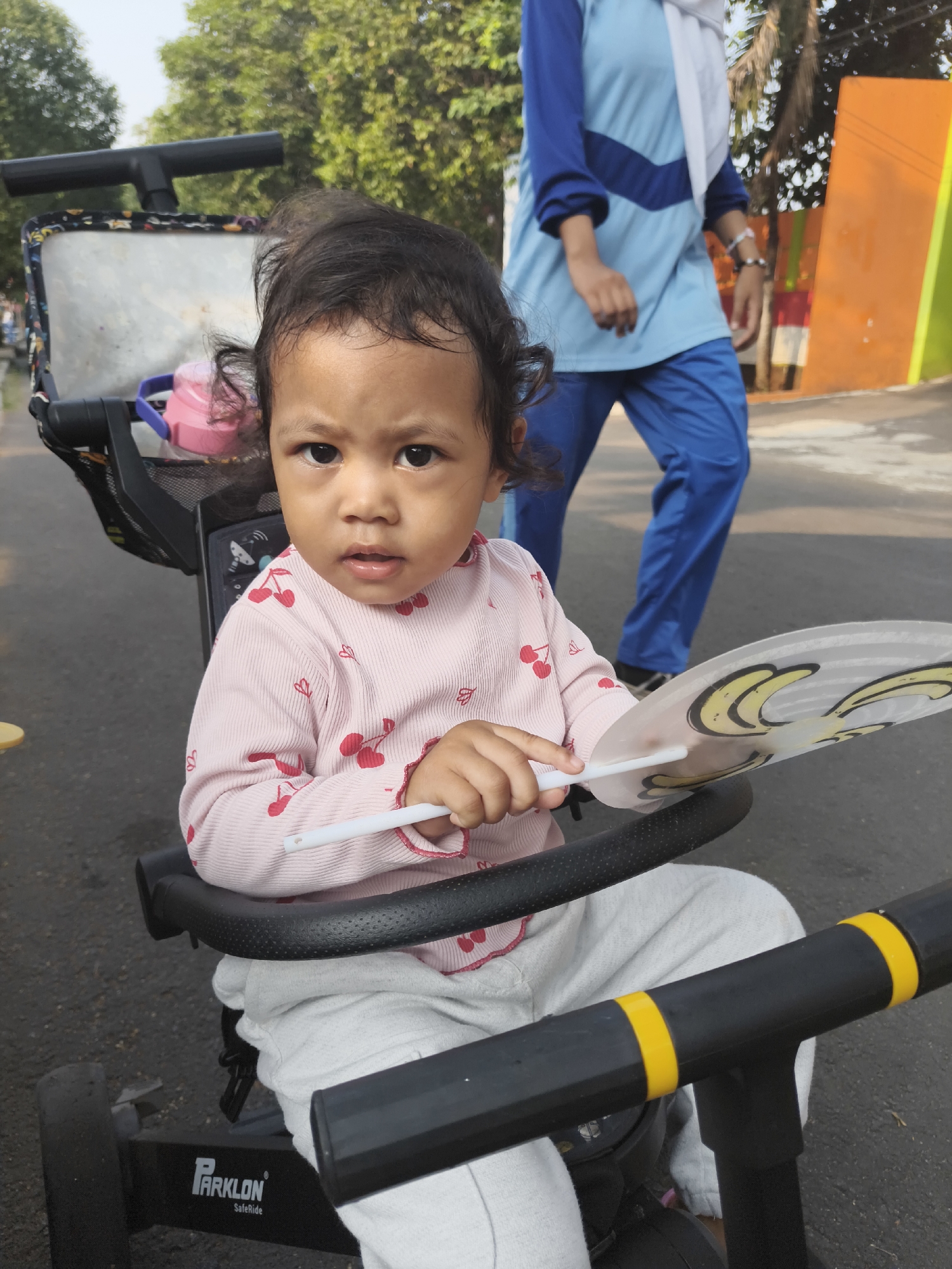In South Jakarta, Indonesia, a young child sits in a black tricycle with a "Parklon SafeRide" logo visible on its frame. The child, wearing a pink long-sleeved shirt with cherry patterns and light gray pants, holds a white fan with a cartoon character design. The tricycle has a black handlebar with yellow accents. In the background, a person in a blue shirt and pants with a white headscarf walks away from the camera, and trees with green foliage line a paved area. An orange and green structure is also visible in the background. The overall lighting suggests it is daytime.