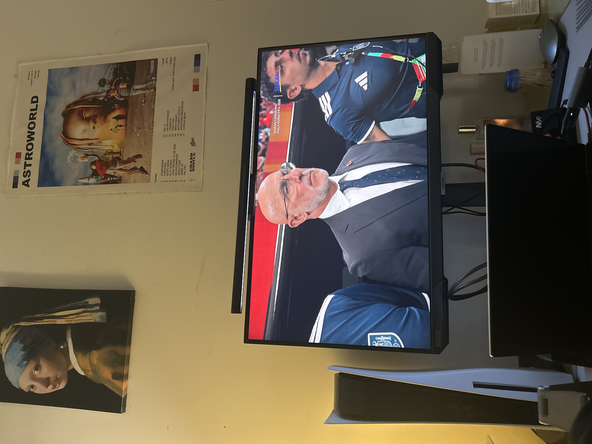 The image depicts a room interior, possibly a bedroom or living area, with the walls painted a pale yellow. On the wall, two posters are visible: one is a reproduction of "Girl with a Pearl Earring" by Johannes Vermeer, and the other is an "ASTROWORLD" album poster. In the foreground, a television screen displays a live broadcast featuring two men. The man on the left is older, wearing glasses, a suit, and a tie. The man on the right is younger, wearing a dark blue athletic jersey with an Adidas logo. The television screen also shows text in Malay, indicating a live broadcast of "PIALA DUNIA FIFA 2026" (FIFA World Cup 2026).
To the left of the television, a white PlayStation 5 console is placed vertically. Below the television, a black laptop is open, and on the desk in front of it, several items are present, including a mouse, a bottle of perfume, and a box of toothpicks. The overall setting suggests a personal entertainment space.
There is no specific information in the image that would indicate the location within Kuala Lumpur, Malaysia.