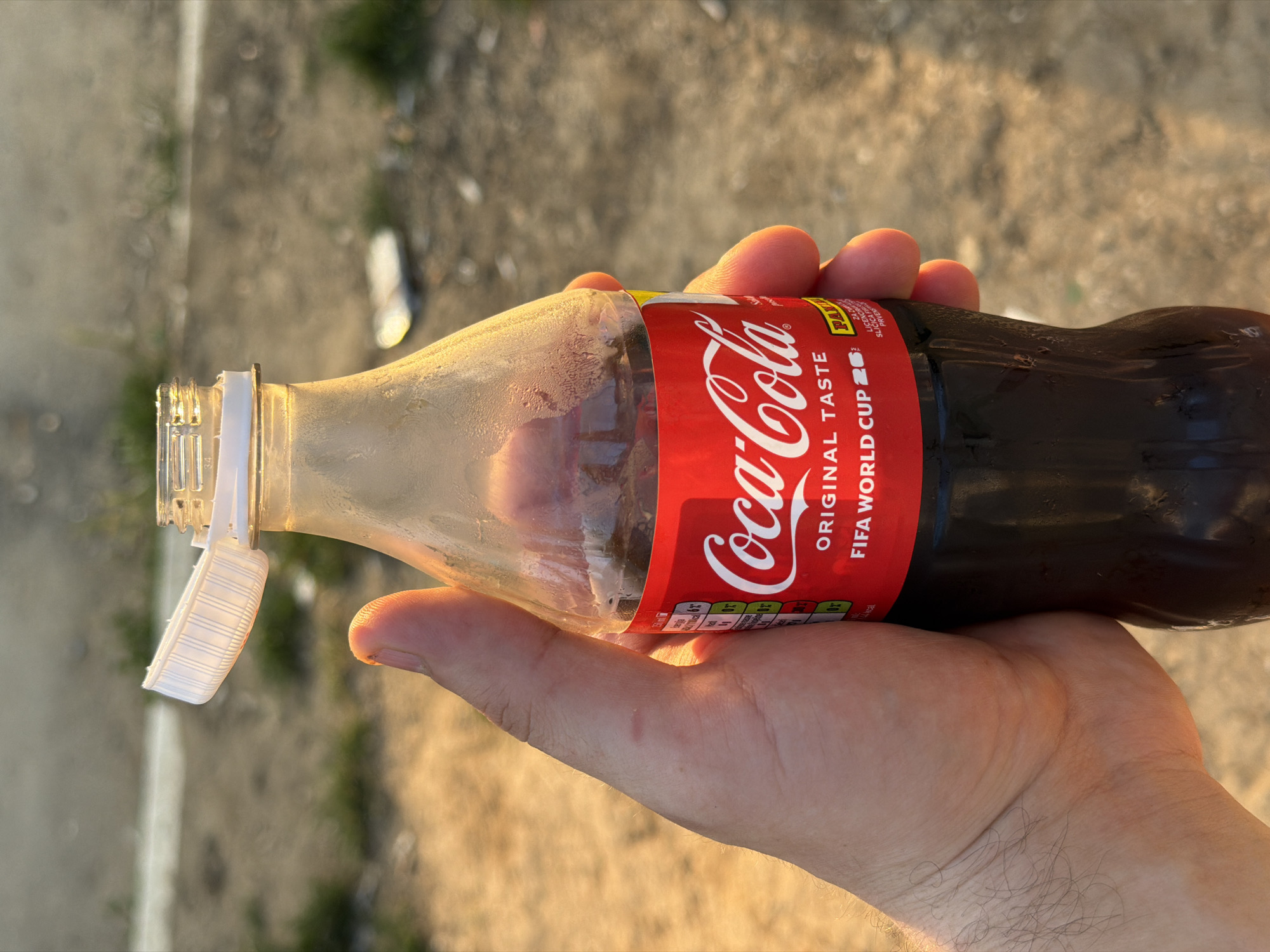 Here is a detailed description of the image:
The image depicts a hand holding a bottle of Coca-Cola. The bottle is partially empty, and the Coca-Cola label is prominently displayed with "ORIGINAL TASTE" and "FIFA WORLD CUP 26". The bottle is translucent, showing the dark liquid inside. The bottle cap is open and attached to the bottle's neck. Condensation is visible on the upper part of the bottle.
The background appears to be an outdoor setting with a blurred, earthy texture, possibly a paved area. The lighting suggests it may be near dusk.