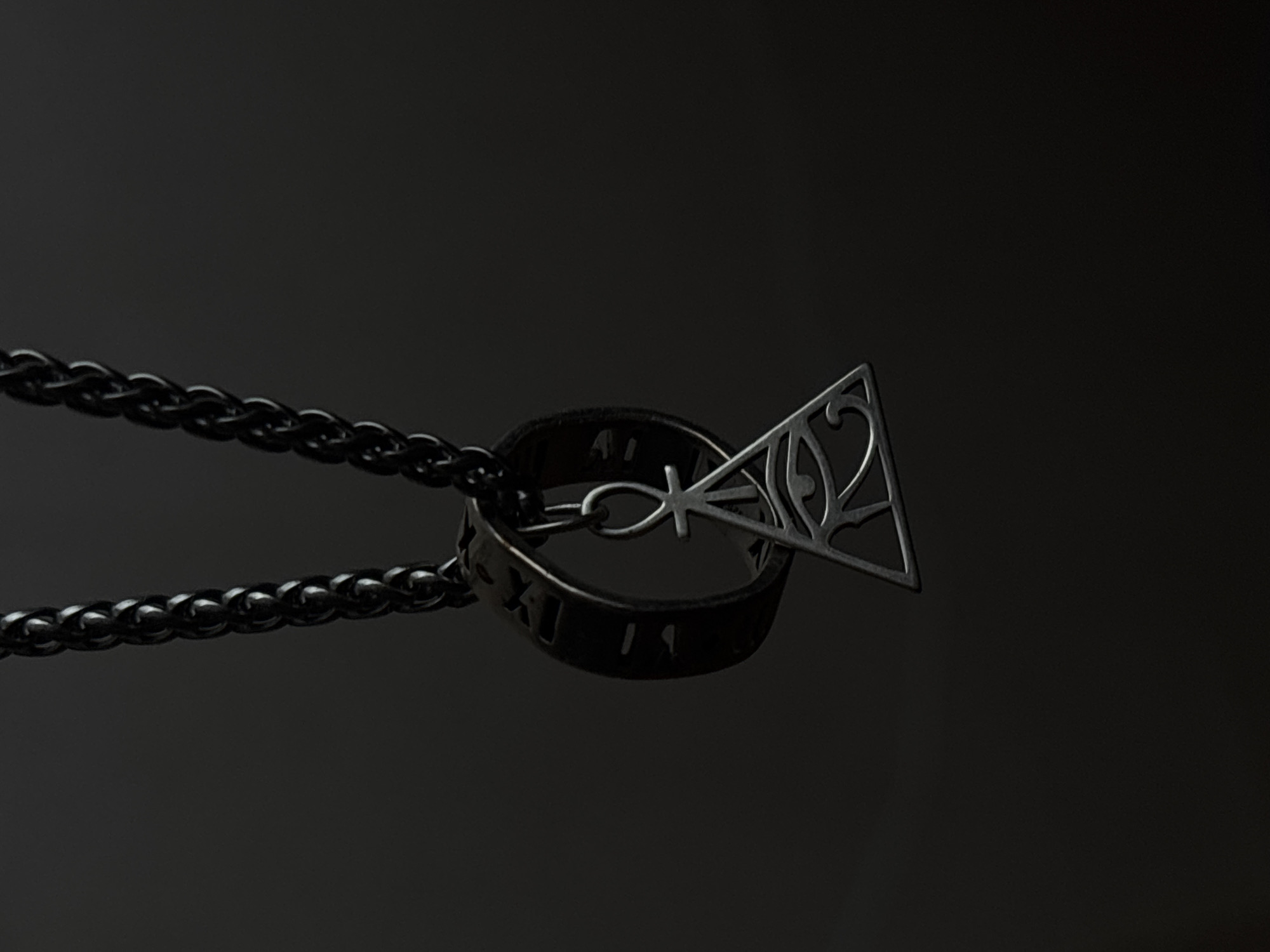 The image displays a close-up of a dark, ornate necklace against a black, reflective background. The necklace features a thick, dark chain leading to a ring charm. This ring is also dark and appears to have carved or etched Roman numerals on its surface, though they are partially obscured and difficult to read clearly. Hanging from the ring is a metallic pendant shaped like a triangle with intricate swirling patterns and an ankh symbol within. The pendant is also dark and has a metallic sheen, suggesting it is made of a dark metal like oxidized silver or gunmetal. The lighting is dramatic, with a spotlight effect highlighting the pendant and ring, casting shadows and creating a sense of depth. The overall aesthetic is mysterious and elegant. No people are visible in the image. The scene appears to be a product shot, likely for jewelry, with the focus entirely on the detailed craftsmanship of the necklace. There is no indication of weather, time of day, or a specific location beyond what is implied by the professional studio setting. The visible text consists of Roman numerals on the ring charm.