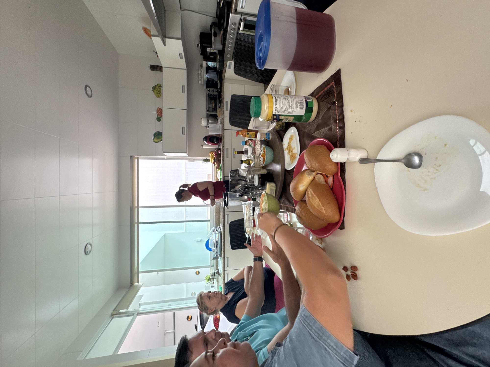 The image depicts an indoor scene within a kitchen in San Bernardino Tlaxcalancingo, Mexico. The kitchen appears well-lit, with white cabinets lining the walls, some decorated with images of fruits and vegetables. There is a large window providing natural light and a view to the outside.
Several people are present. One person is seen in the background near the stove, wearing a red shirt. Three people are seated at a table closer to the camera.
The table is covered with food items. A plate with food residue and a spoon is in the foreground. A red bowl containing bread rolls is also visible. Other items on the table include a pitcher filled with a red liquid, a jar of parmesan cheese, and other containers. Scattered on the table are a few pieces of small, brown food.