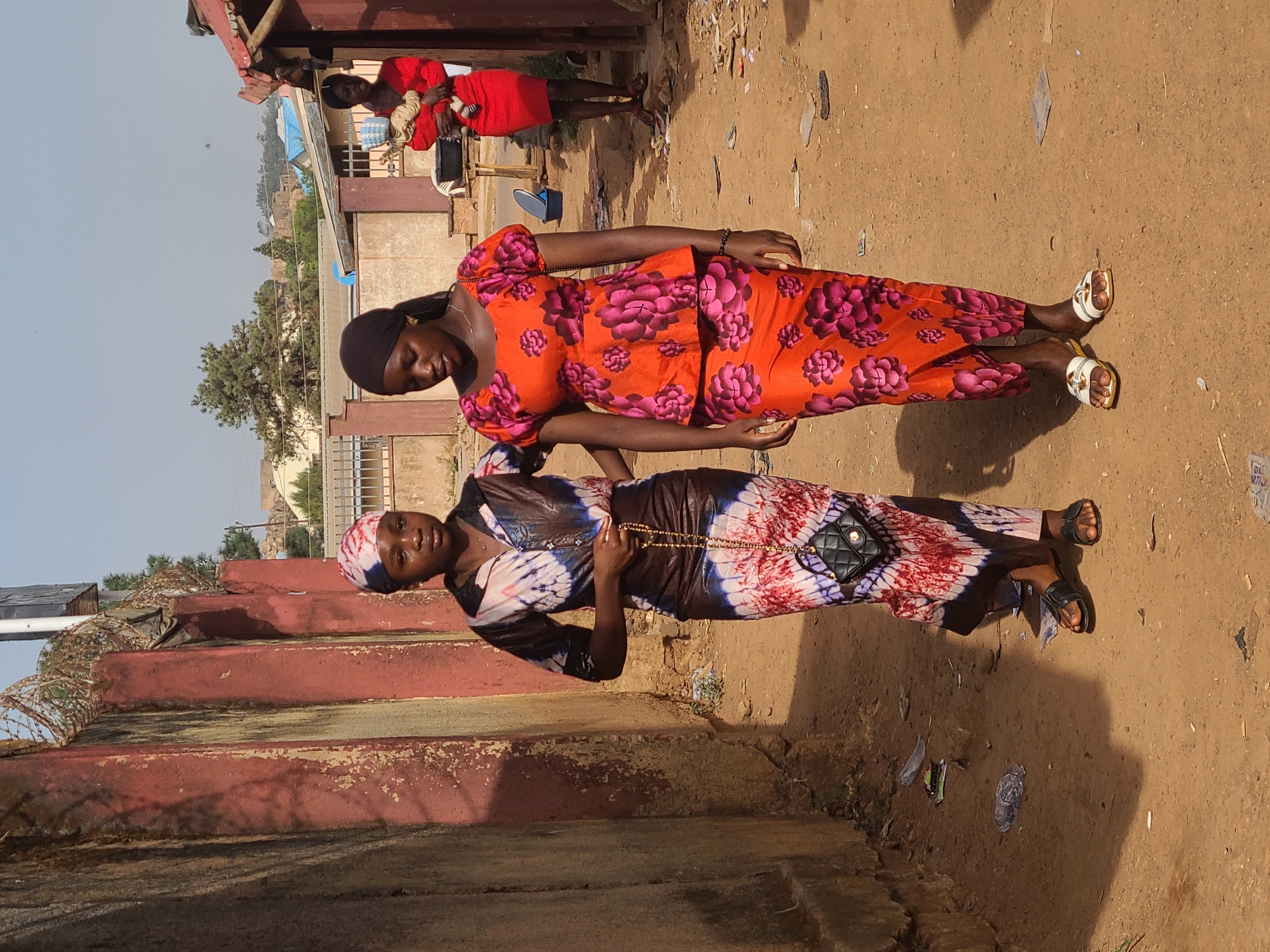 The image shows two women standing on a dirt road next to a red wall. One woman is wearing an orange and pink floral patterned outfit, while the other is wearing a tie-dye dress with a black purse. A third woman in a red dress is visible in the background, holding a baby. The location appears to be in Jos, Nigeria, indicated by the architecture, clothing styles, and general environment. There is trash on the ground, and barbed wire is visible on top of the wall. The lighting suggests it is daytime.