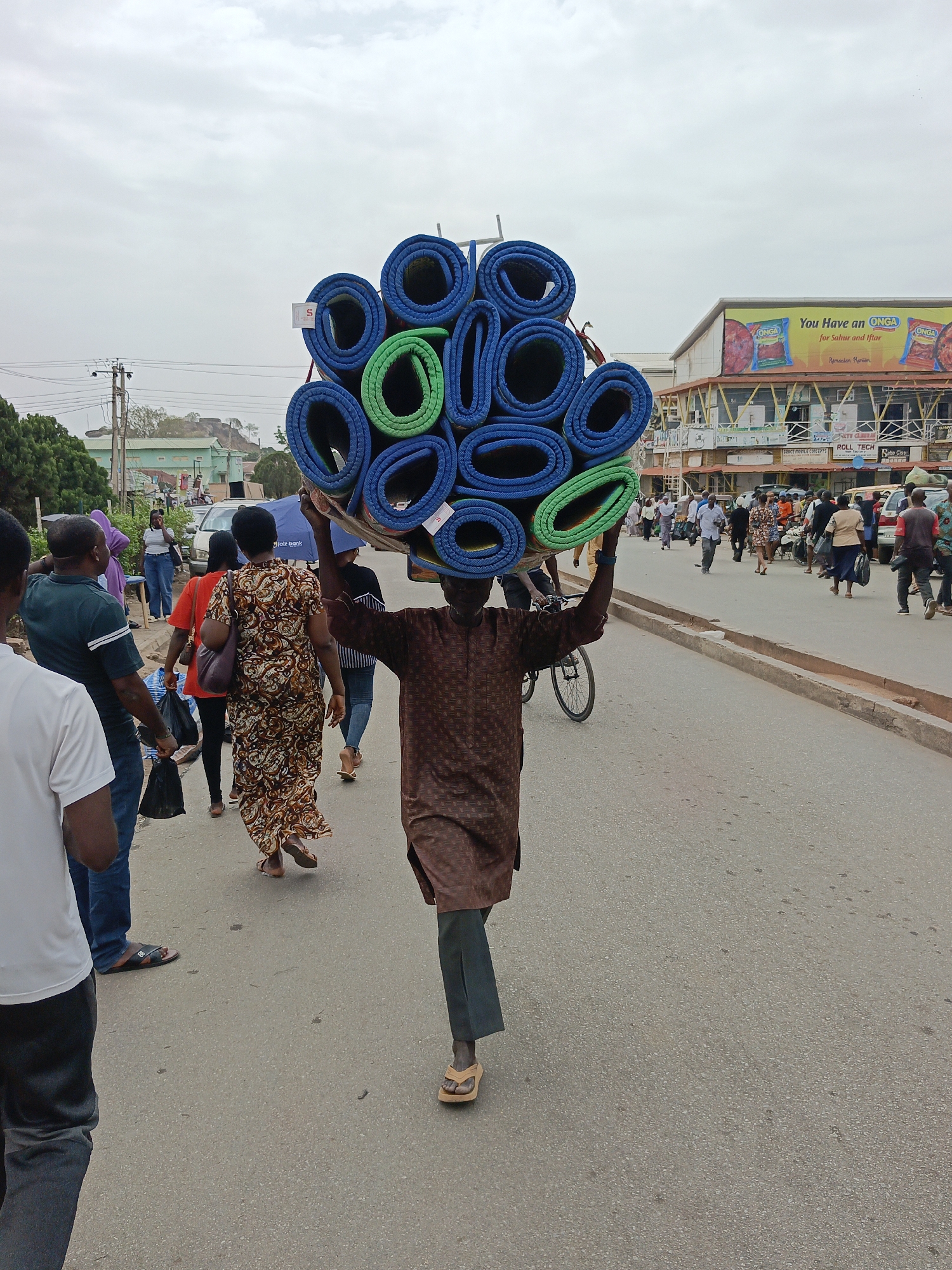 A wide, eye-level shot captures a man carrying a large bundle of rolled-up exercise mats on his head. The mats are predominantly blue, with some green ones interspersed. He is walking towards the viewer on a paved street, wearing a long-sleeved brown shirt with a patterned design and dark pants. His feet are clad in flip-flops.
Around him, other pedestrians walk in both directions, some facing the camera and others with their backs to it. A bicycle is visible in the background, near the man. On the right side of the frame, a sidewalk with more people is evident. A building with several businesses and an advertisement featuring the word "ONGA" can be seen in the background.
The sky above is overcast and pale. Utility poles and wires are visible in the upper left corner of the shot. The scene is a busy street with people engaged in various activities.