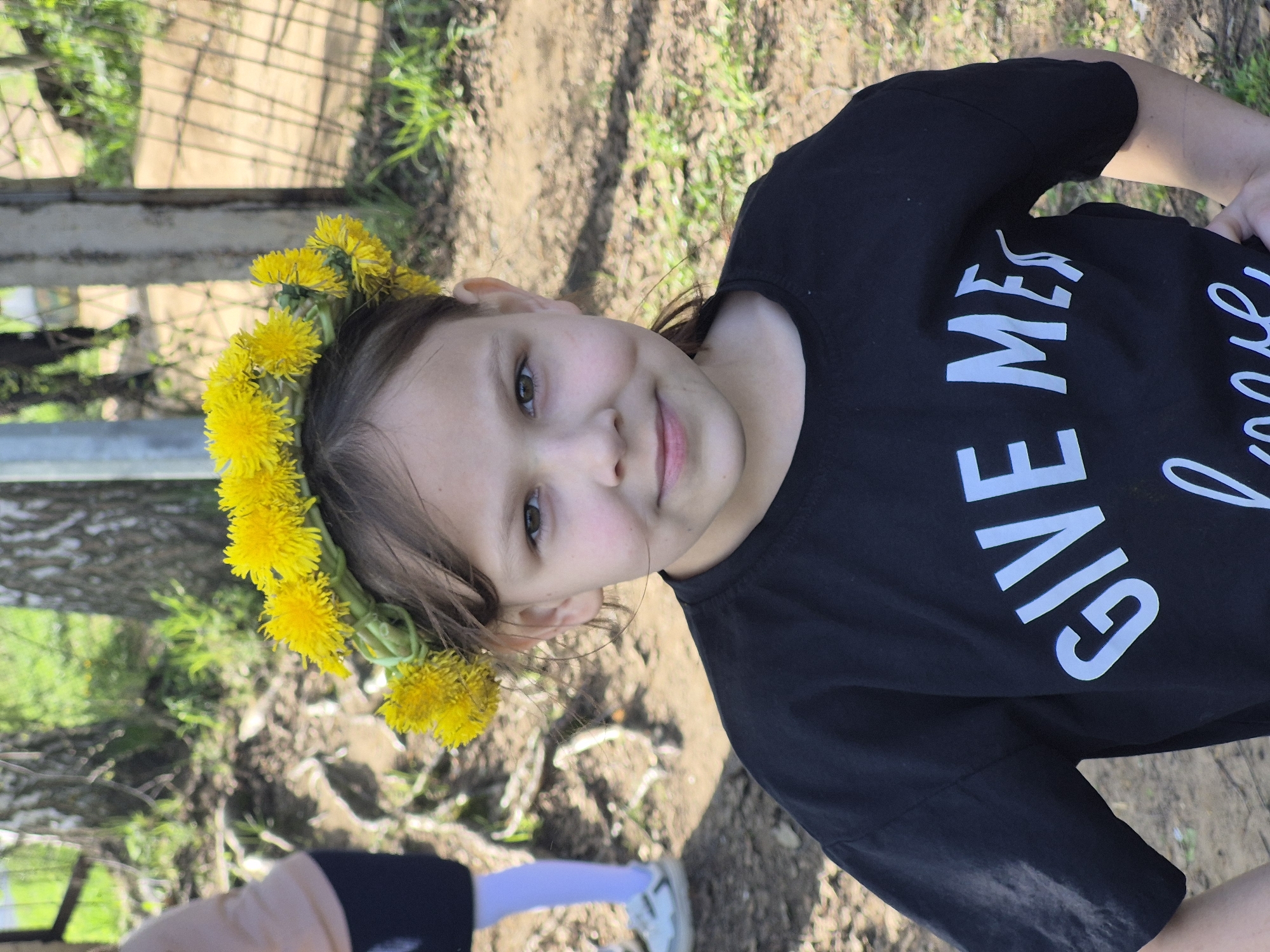 A young girl is outdoors wearing a dandelion crown. She is wearing a black t-shirt with the white text "GIVE ME". The setting appears to be a park or yard, with dirt and vegetation visible. Part of a fence is visible to the left of the girl.
