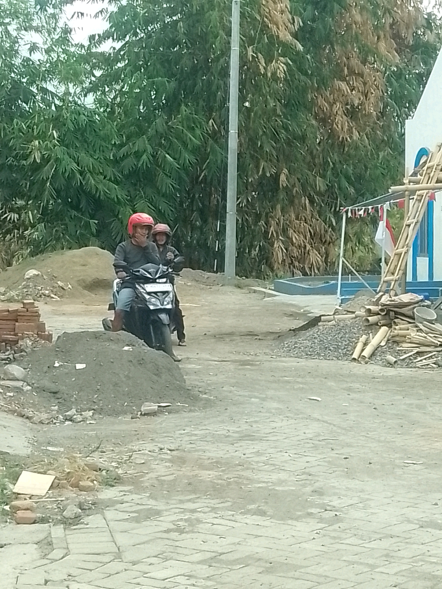 Two individuals wearing helmets are riding a black scooter on a dirt road in Jember, Indonesia. The person in front is wearing a red helmet and a dark long-sleeved shirt, while the person behind is wearing a lighter helmet and a dark jacket. They are positioned behind a pile of gray dirt and in front of a large bamboo grove. To the right of the bamboo grove, a white building with blue trim and a ladder made of bamboo is visible, along with some decorative flags. The ground is a mix of dirt, gravel, and paved brickwork. The scene appears to be a construction site or an area undergoing development.