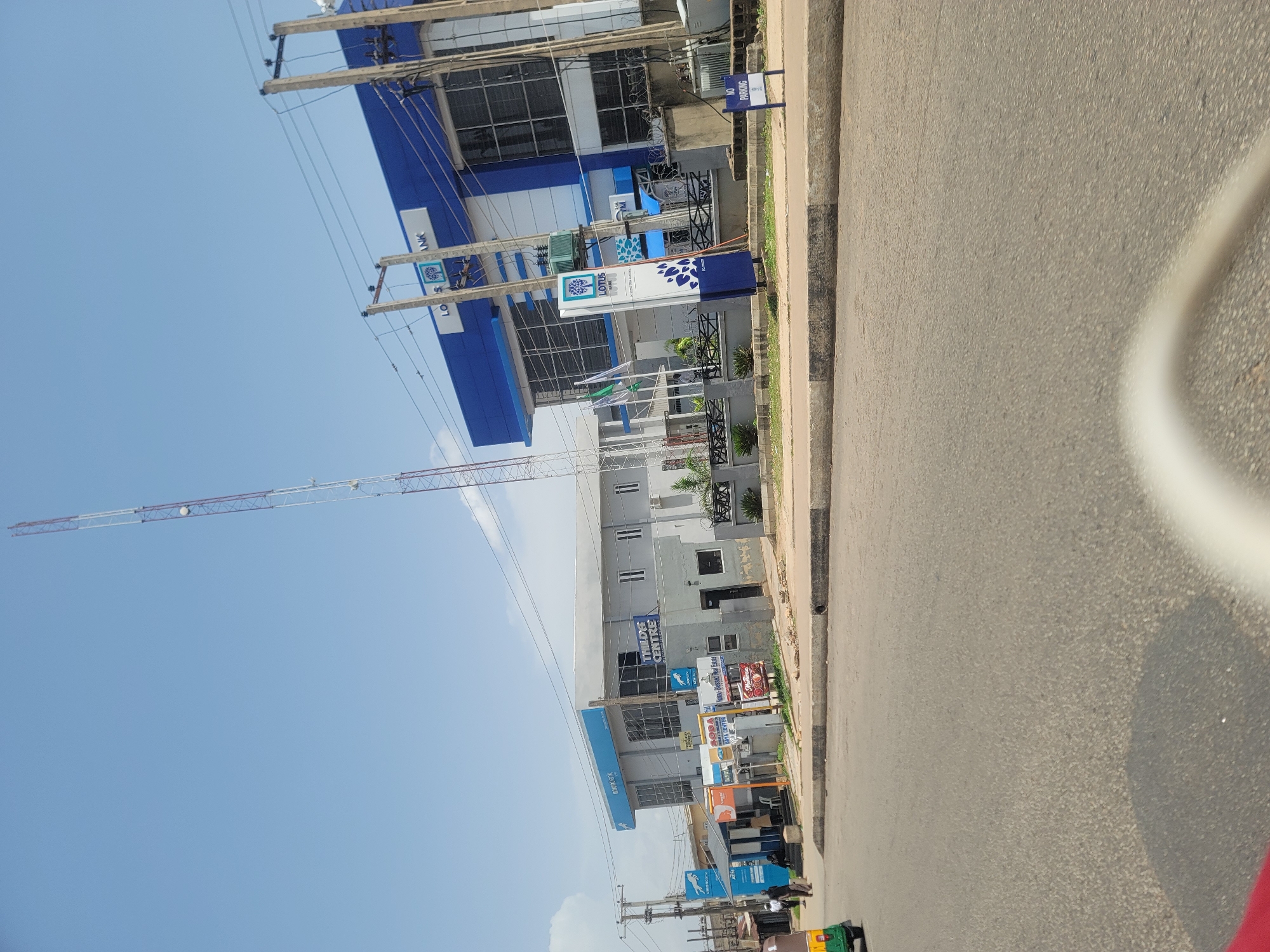 The image captures a street scene in Jos, Nigeria. A building with a prominent blue and white facade, identified by signage as "UNIONBANK" and "LOTUS BANK," dominates the left side of the frame. Adjacent to it is another building with multiple windows and the text "THILDEG CENTRE" visible. Several other signs are present, including "SOPA," "Katau-Blessed Real Estate," and a "NO PARKING" sign.
A tall construction crane extends into the clear blue sky from the left side of the image, suggesting ongoing development or construction in the area. Overhead power lines crisscross the scene, supported by wooden poles. The right side of the image shows a paved road with a gray curb. The overall impression is of a busy urban environment, likely during the daytime given the bright sky and shadows, though no people are clearly depicted engaging in any specific activity. The weather appears to be clear and sunny.