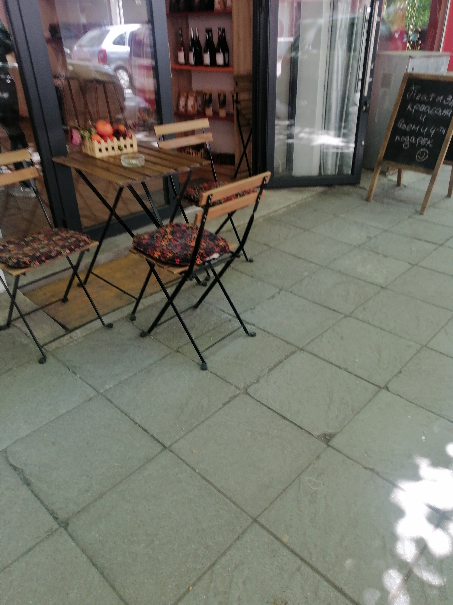 The image depicts an outdoor seating area of a café or small shop. Two chairs and a small table are arranged on a paved surface consisting of gray square tiles. A basket with fruits and what appears to be an ashtray is on the table. The chairs have cushions with a floral pattern. Behind the seating area is a storefront with a glass door and window, through which products like bottles of wine can be seen. A car is visible through the glass in the background. To the right, a chalkboard stands, displaying text in what appears to be Bulgarian, with an advertisement of a "Treat" and a "gift" in Bulgarian. The scene is likely in Sofia, Bulgaria, judging by the style of the storefront and the writing on the chalkboard.