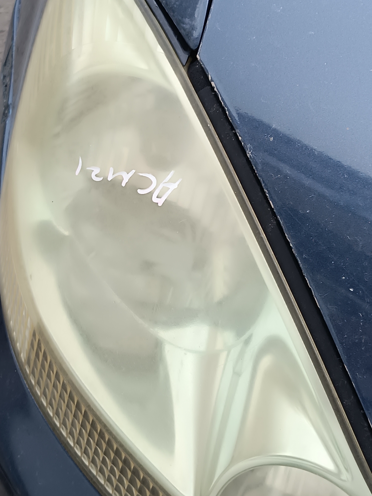The image shows a close-up view of a faded yellow car headlight with white writing on it. The writing appears to be "HCM21". The headlight is attached to the dark blue body of the car. The scene is outdoors, but no specific city or location details are visible. The lighting suggests it is daytime, but there are no other environmental clues.