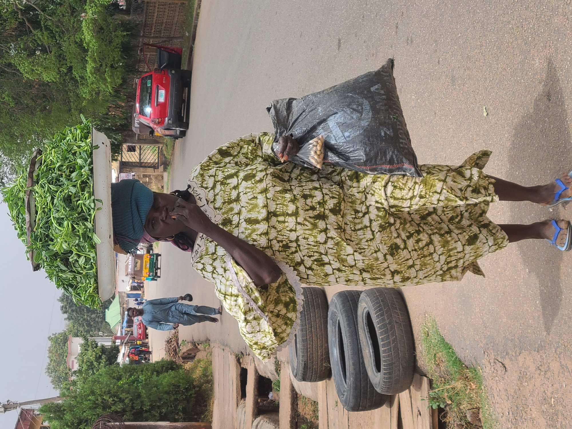 A woman walks along a road in Jos, Nigeria. She balances a large tray of leafy green vegetables on her head. She carries a dark plastic bag in her other arm. She wears a green and white patterned dress and blue sandals. Three used tires are visible along the side of the road near a storm drain. A red car and other vehicles are in the background. A man walks on the other side of the road.