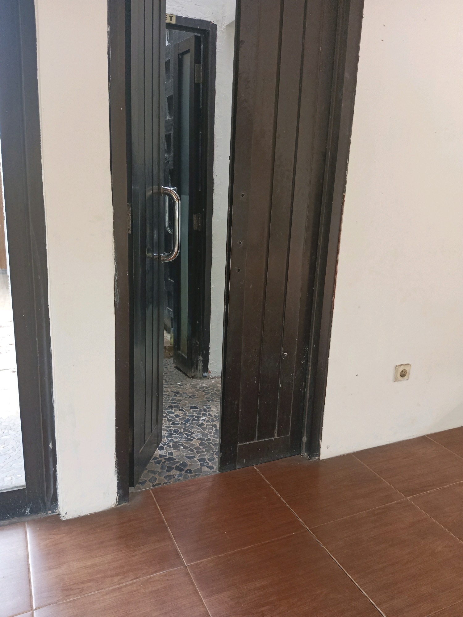 The image depicts an interior view of a hallway or entryway, with multiple doors visible. In the foreground, a brown wooden door stands open to the right, revealing its vertical paneling. To the left, another dark-colored door frame is visible, with the edge of a glass-paneled door in the far left.
The primary focus is the open dark wooden door, which has a polished silver handle. Through this doorway, another dark wooden door is visible, slightly ajar. Beyond that, a third dark wooden door is also partially open. The floor visible through the first open doorway is covered with a mosaic of small, dark grey and black stones.
The walls are painted white. On the wall to the right, at approximately eye level, there is a white electrical outlet. The floor in the foreground is tiled with large, square, brown tiles.
The scene suggests an indoor space, possibly a commercial establishment or a residential building with multiple rooms or units. Given the style of the doors and tiling, it is plausible that this scene is located in South Tangerang, Indonesia, a region known for its developing infrastructure and mixed architectural styles. There are no people or animals present in the image. The lighting appears to be artificial, with shadows cast by the doors.