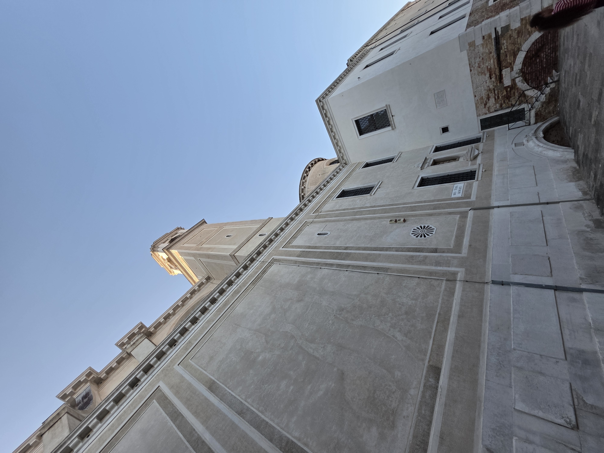 An exterior, low-angle shot captures a section of a large, light-colored building in Venice, Italy, under a clear blue sky. The building features a tall, square tower to the right of center, with a domed structure visible behind it. The facade of the main building is textured and decorated with relief lines and a circular, star-like emblem. Several windows are visible, some with decorative grilles. The lower portion of the building has large arched openings and is made of lighter-colored stone. To the right, a smaller section of the building is visible, with exposed red brickwork forming arched recesses. A person wearing a striped shirt is partially visible on the far right, walking away from the camera. The ground in front of the building is paved with stone. A street sign reading "RIO TERRÀ DEI DESCRATI" is mounted on the building.