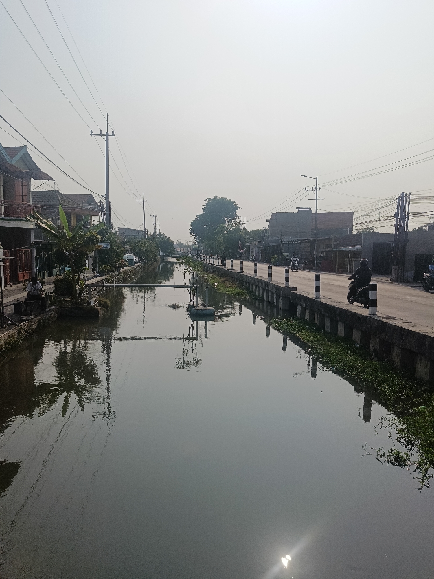 The image depicts a canal running through the town of Wedi, Indonesia. The canal is flanked by a paved road on the right and a sidewalk and buildings on the left. The water in the canal is still and reflects the surrounding environment, including buildings, trees, and utility poles. Several utility poles with wires run along the road and the canal.
On the road, there are several motorcycles, with riders in helmets, indicating ongoing traffic. A person is seated on a bench on the left sidewalk, near some potted plants and a small tree. The buildings on the left appear to be residential or commercial, with varied architectural styles and roofs. The environment is hazy, suggesting either smog or a bright, overcast day. The canal itself is bordered by a concrete embankment with black and white striped posts along the road. Green vegetation is visible along the edges of the canal and on the embankment. The sun is visible as a bright glare on the surface of the water, suggesting it is daytime.