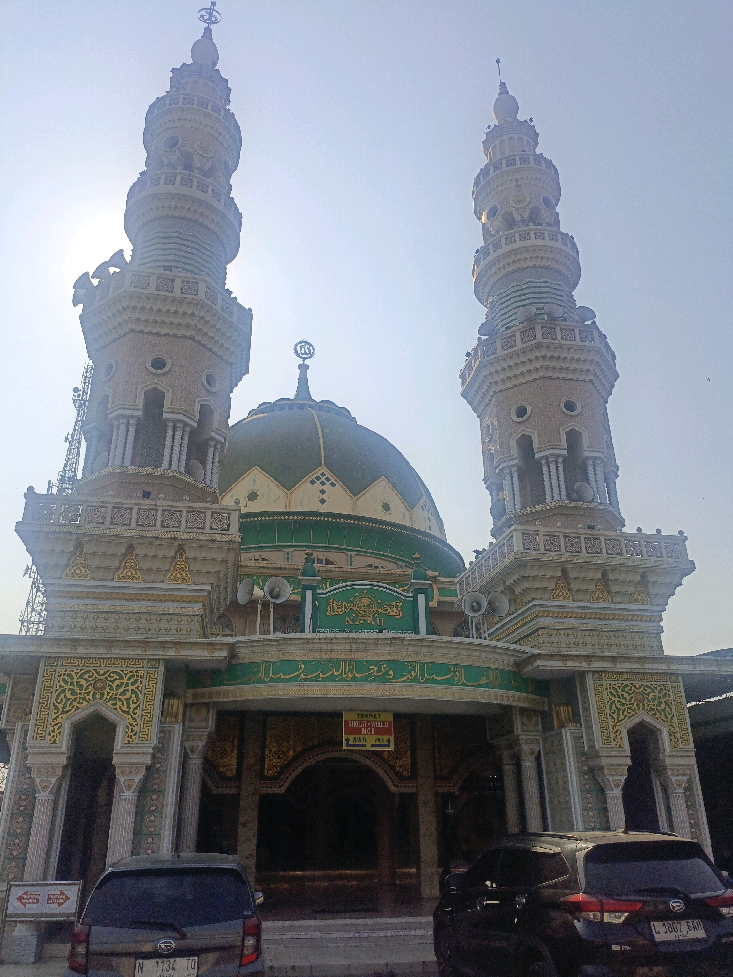 The image captures the facade of a mosque, characterized by its ornate architecture. The mosque features two prominent minarets flanking a central dome, all adorned with intricate patterns and decorative elements. The minarets are tall and slender, with multiple tiers and balconies, topped by spires. The dome is green and rounded, with a crescent moon and star emblem at its apex. Below the dome, the mosque's entrance is elaborate, with arched doorways and decorative panels featuring Islamic calligraphy and geometric designs. Several loudspeakers are visible on the exterior, likely for calls to prayer. Two cars are parked in front of the mosque, one in the foreground on the left and another to the right. Signs are visible near the entrance, one of which appears to indicate directions for prayer facilities. The sky is bright and clear, suggesting a sunny day. The overall impression is of a well-maintained and visually striking religious building, likely in a populated area given the presence of cars and the urban infrastructure implied by the mobile towers in the background. The presence of the cars, the architectural style, and the visible text on the signs, when combined with the location provided, strongly suggest this is the Masjid Agung Al-Ukhuwah, located in Gempol, Indonesia.