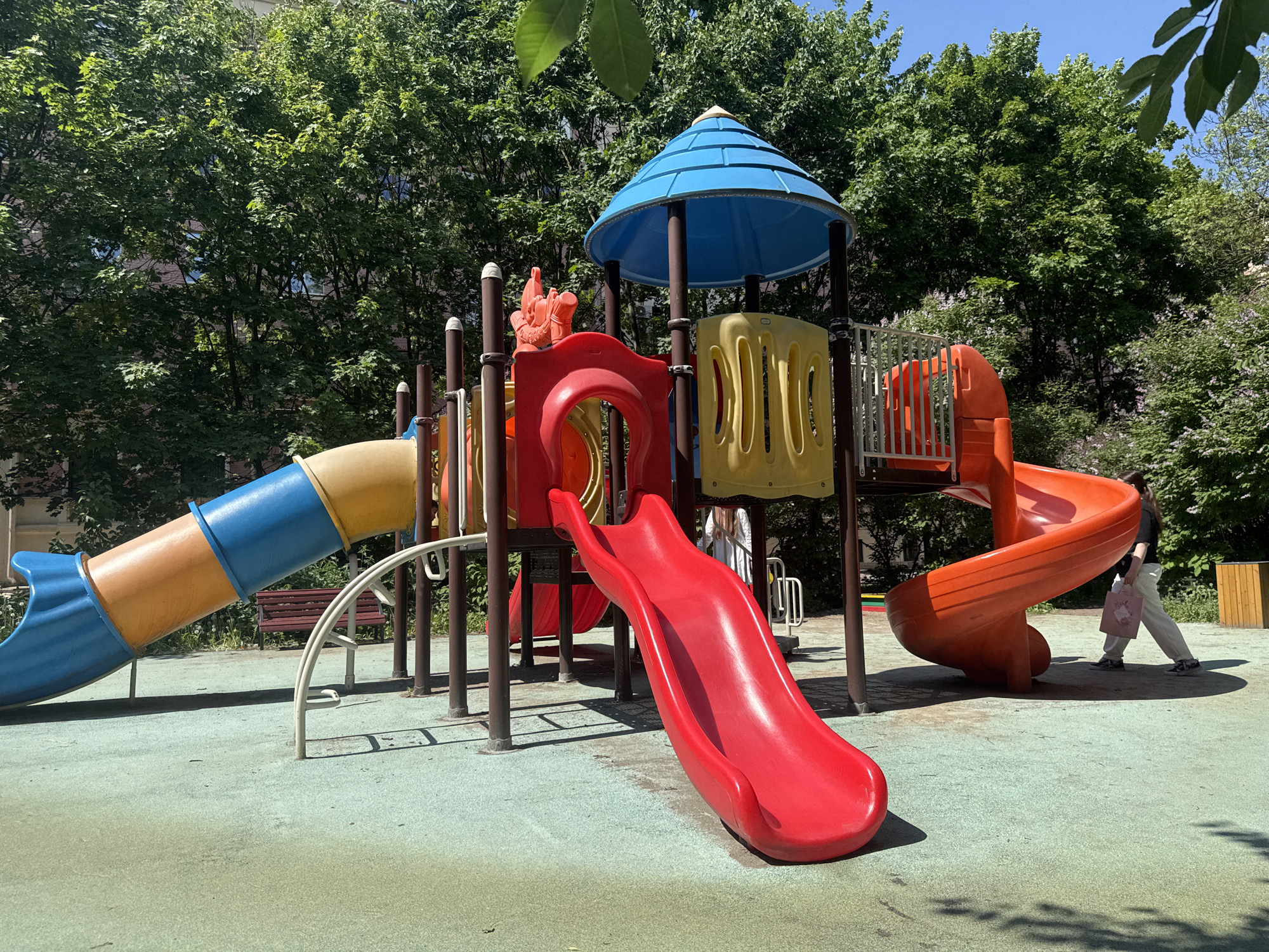 This image depicts a vibrant, multi-colored children's playground located in Saint Petersburg, Russia, under bright sunshine. The scene is set outdoors, surrounded by lush green trees and foliage, suggesting a clear spring or summer day. The ground surface is a soft, textured light green and blue rubber, typical of modern play areas, with some visible wear.
The central element is a large, composite playground structure built from brightly colored plastics and metal. It features three prominent slides: a red, straight slide in the foreground, a blue and yellow enclosed tube slide curving downwards on the left, and an orange spiral slide on the right. The structure also includes platforms, stairs, climbing components, and is topped with a distinct blue conical roof.
Two individuals are visible. On the far right, a person wearing a dark top and light-colored pants is walking away from the playground, carrying a light pink bag adorned with a graphic. Another person is partially obscured behind the central structure and the red slide, only their lower body and part of their torso visible in light clothing. Neither individual appears to be actively playing on the equipment.
Strong sunlight casts distinct shadows from the playground structure and trees across the ground. A wooden park bench can be seen in the background under the tube slide, and a wooden trash bin is partially visible on the far right. There is no discernible text visible on any objects within the frame. The overall impression is that of a quiet moment at a colorful and well-equipped outdoor play space on a sunny day.